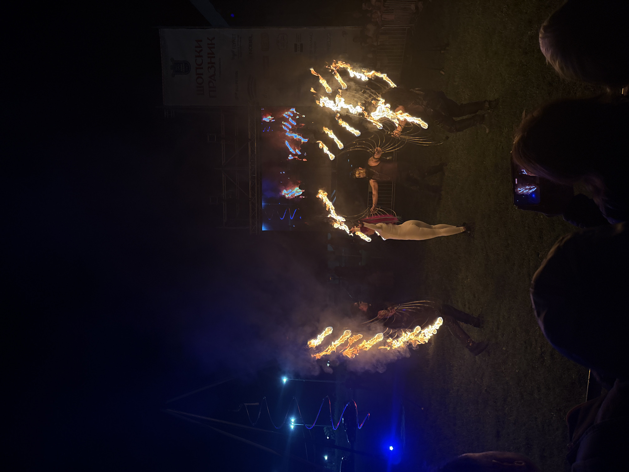 This nighttime image captures a fire performance during an outdoor festival in Elin Pelin, Bulgaria. In the foreground, the silhouetted heads of several spectators are visible, with one individual on the lower right filming the event horizontally with a smartphone, its screen showing a bright, blurry reflection of the flames.
The central focus is on four performers on a grassy area. From left to right: a performer in dark attire creates a vertical line of intense flames and smoke with a fire prop, facing right. To their right, a second performer, dressed in a light-colored, form-fitting suit with pink hair, holds a flaming hoop, facing towards the right. A third performer, with dark hair and wearing dark costume elements, stands center-right, manipulating several flaming hoops to create a broad fan-like display of fire. The fourth performer, positioned on the far right and also in dark clothing, mirrors this action with multiple flaming hoops, facing towards the left. The ground is green grass, illuminated by the fire and ambient blue stage lighting.
In the mid-ground on the left, a tall, slender structure with a glowing red and blue spiral design ascends, with blue spotlights at its base. In the background on the right, a large white banner is prominently displayed, featuring the Cyrillic text "ШОПСКИ ПРАЗНИК" (Shopska Feast) at the top, along with smaller sponsor logos like "FLYNET" and "NOBEL." Below the banner, a large screen exhibits abstract, vibrant light patterns. Scaffolding structures support the banner and screen. Beyond the performance area, behind a metal barrier, a dimly lit crowd of additional spectators can be discerned. The sky above is dark, indicative of nighttime.