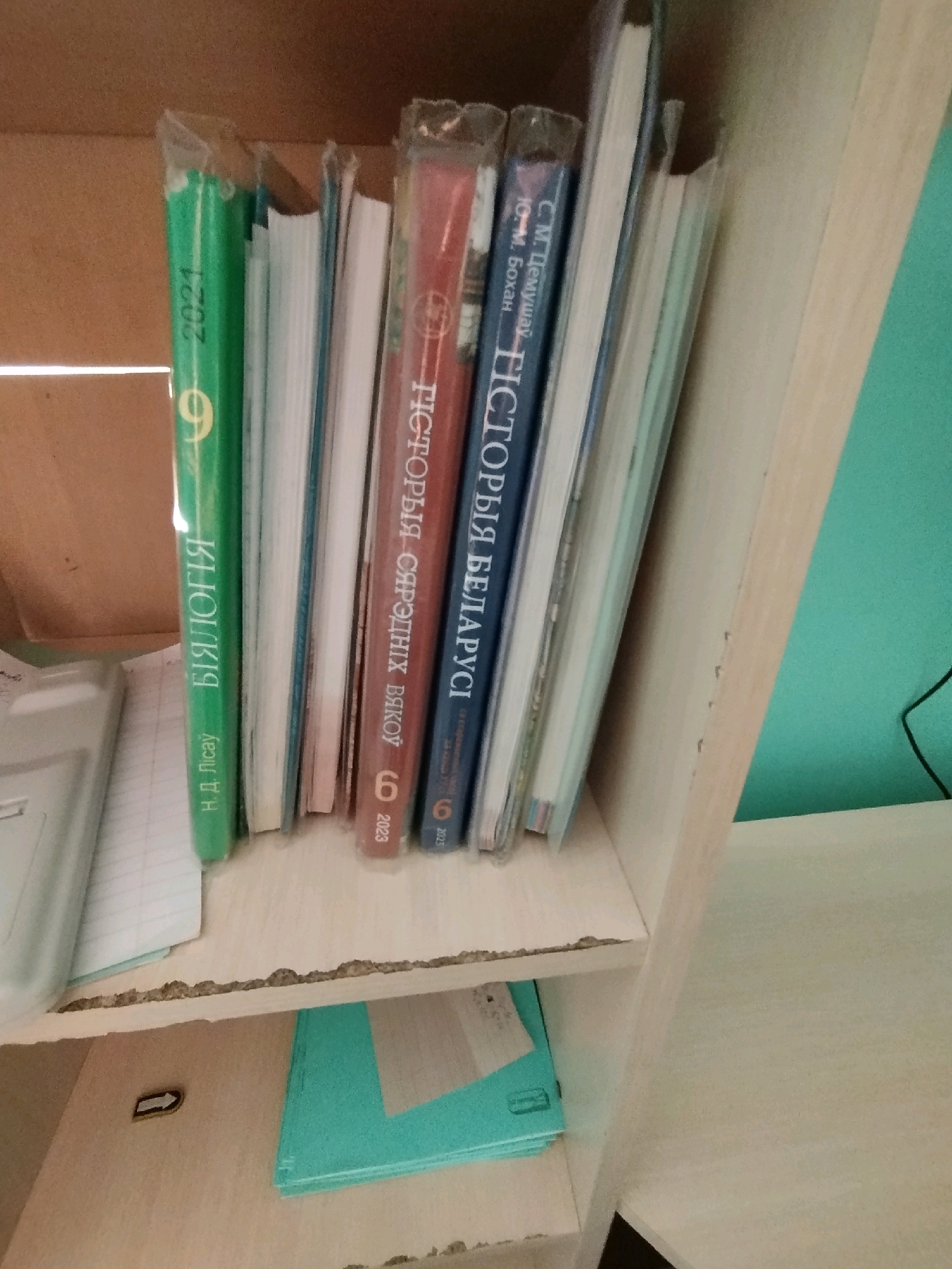 On a wooden shelf, a collection of books is arranged. The spines of the books are visible, with some titles in Cyrillic script. One green book has "БІЯЛОГІЯ" and "Н.Д. Літоўскі" printed on it, along with "2021" and a "9". Another book, red, has "ГІСТОРЫЯ" and "СЯРОДНІХ" visible, with "2023". A blue book also shows "ГІСТОРЫЯ" and "БЕЛАРУСІ", with "2023". There are several other books with white and blue spines. To the left of the books, a white, partially visible object appears to be a notebook or folder. Below the shelf, a turquoise folder is visible. The background consists of a brown surface and a light-colored wall. This scene is located in the village of Krivaselski, within the Krivaselski village council, Belarus.