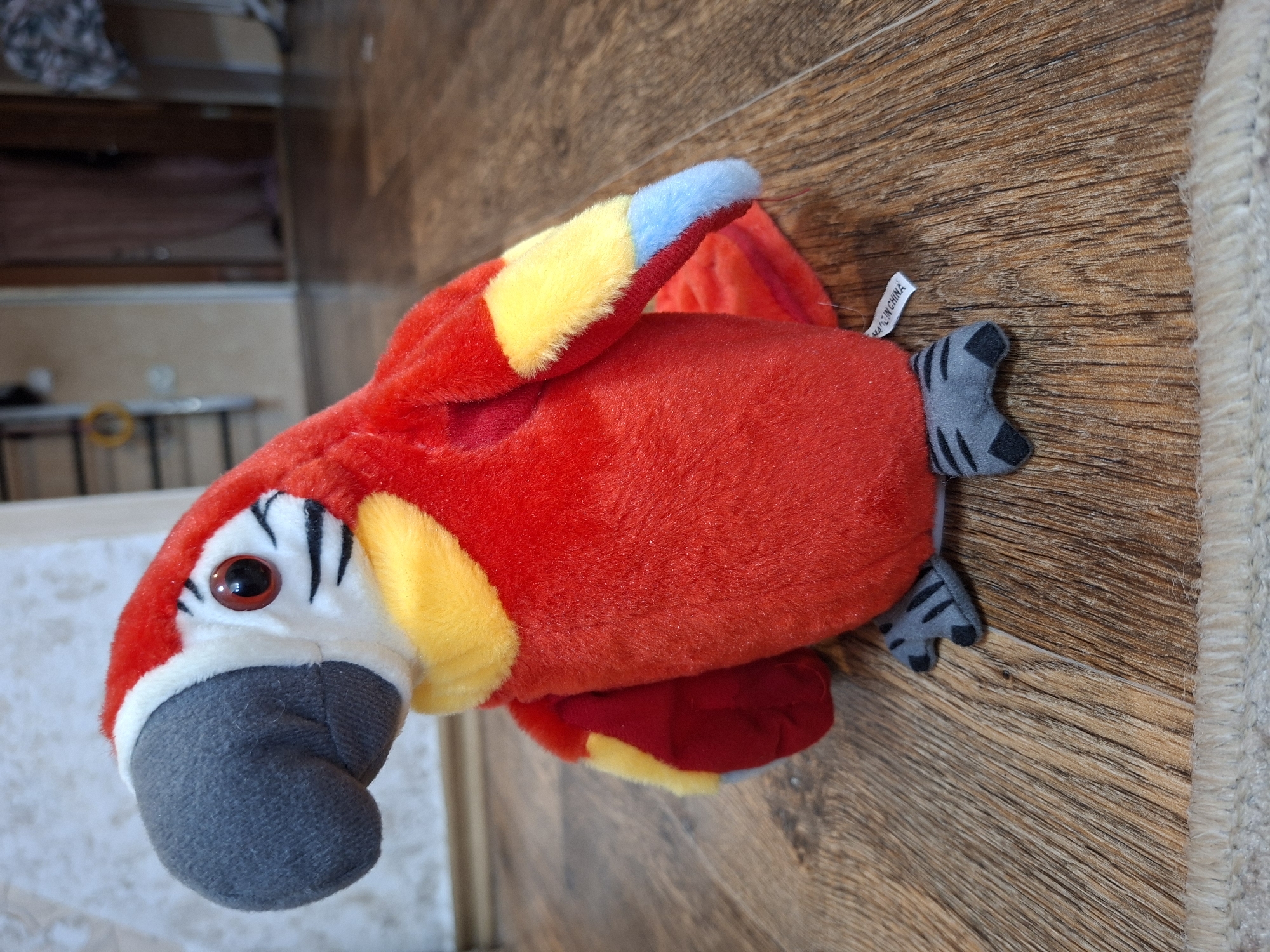 A plush toy parrot with red, yellow, blue, and gray coloring is sitting on a wooden floor. The parrot is positioned facing left and is slightly angled towards the viewer. Its beak is gray, and its eyes are red with black pupils. Yellow and white feathers surround its neck, and red and yellow feathers adorn its wings. The parrot's feet are gray with black stripes. A small white tag that reads "MADE IN CHINA" is attached to its body. The background shows a wall with a light-colored texture, a portion of a door frame, and some blurred objects. The floor is made of wood planks with visible grain. This scene is located in Gorkovskiy, Russia.