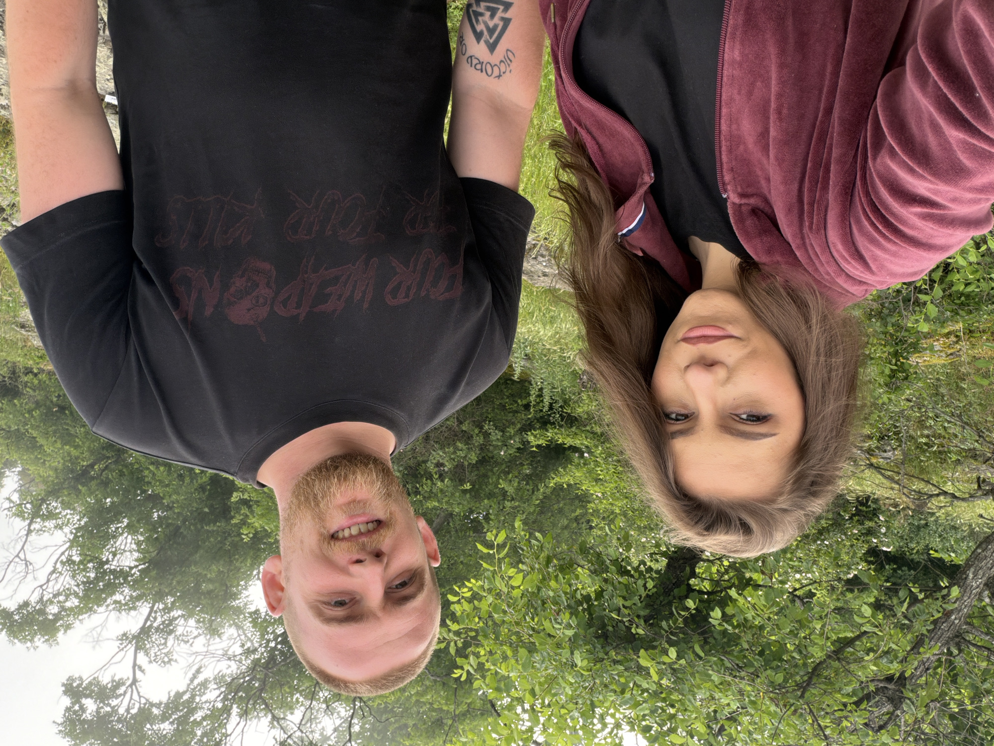 An outdoor portrait shot captures two individuals, a man and a woman, positioned with their heads at the bottom of the frame and their bodies extending upwards, suggesting the image is rotated. They are set against a vibrant green backdrop of dense trees and foliage under what appears to be an overcast sky.
The man, on the left, has a light brown beard and short hair, and is smiling broadly, revealing his teeth. He wears a black t-shirt adorned with dark red text and a skull graphic. The text on his shirt, though partially obscured and upside down, reads "WEAPONS FOR OUR KILLS". On his right forearm, a prominent black tattoo features a Valknut symbol and the phrase "VICTORY OR DEATH".
The woman, on the right, has long brown hair and a neutral expression, looking directly at the viewer. She is dressed in a black top covered by a rich burgundy or maroon-colored zippered jacket with a soft, possibly velvet, texture.
The setting is a natural environment, likely a forest or park, indicated by the abundant green leaves and tree branches filling the background. The lighting is soft and diffused, characteristic of a cloudy daytime.