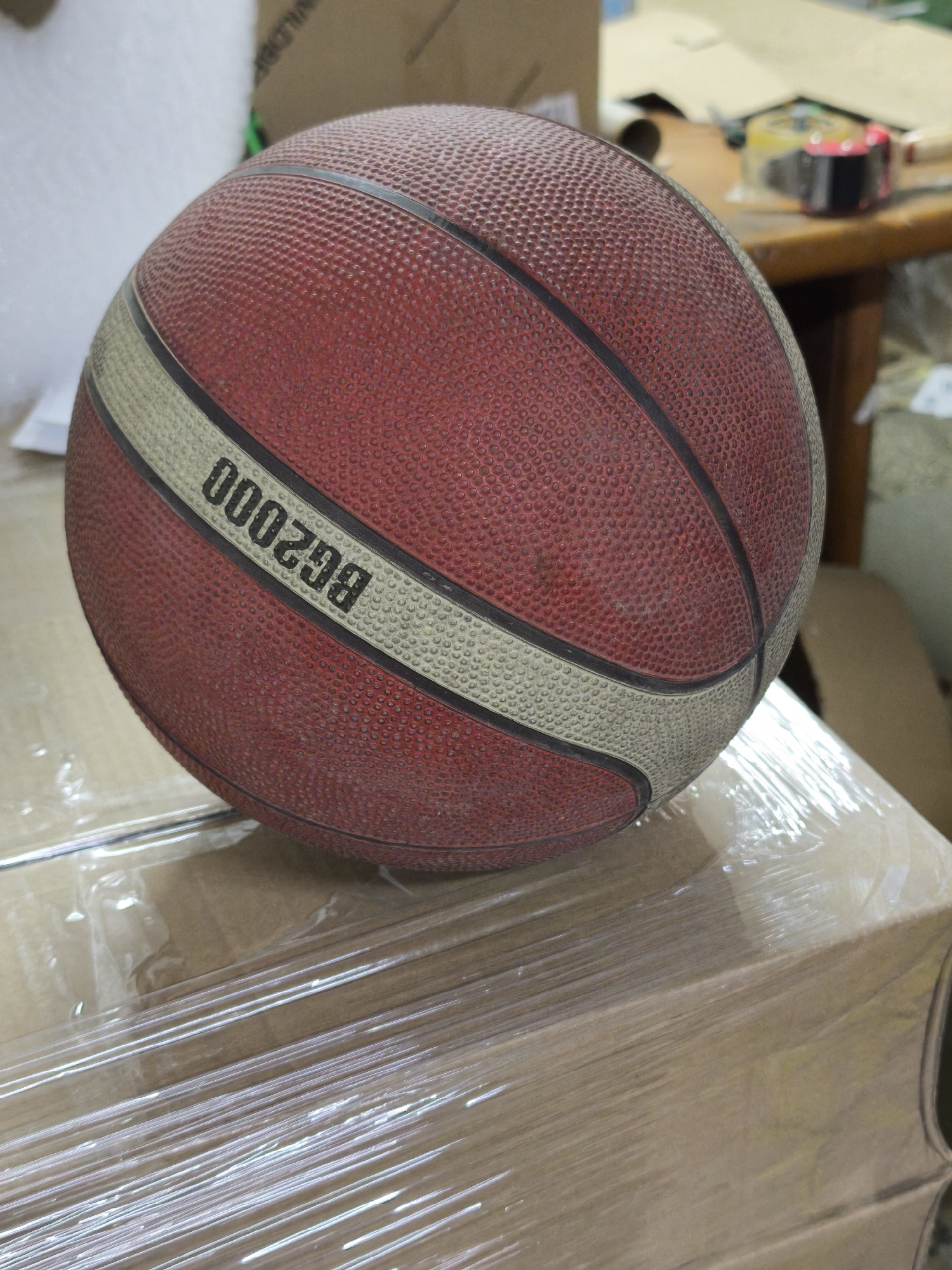 A red and white basketball with the printed text "BG2000" is placed on a box wrapped in clear plastic. The background shows a blurred room with various objects including boxes and a wooden table. The location is not possible to determine from the given visual information.
