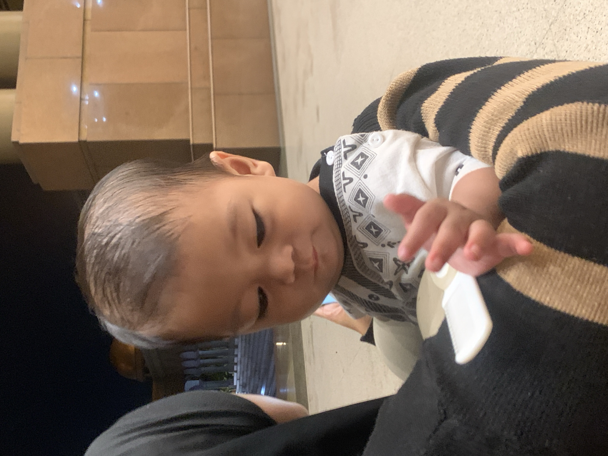 The image features a close-up, slightly tilted view of a baby or young toddler being held by an adult. The baby, positioned prominently in the left half of the frame, has dark, slicked-back hair and a soft, downward gaze, suggesting curiosity or focus on something below. Their right ear is visible.
The child is wearing a white short-sleeved top adorned with a black and white geometric pattern, possibly a traditional or tribal design, particularly visible on the front and shoulders. Their left hand is gently touching a small, white rectangular tag or label, which appears to be attached to the adult's clothing. The adult, whose face is not visible, is wearing a garment with prominent black and tan horizontal stripes on the right side, likely a sweater or long-sleeved top. A plain black fabric is also visible in the lower left, possibly part of the adult's attire.
The background is softly lit, indicating an indoor setting or a covered outdoor area at night or in the evening. Light-colored, possibly tiled or stone, architectural elements are visible in the upper background, with reflections from artificial light sources creating soft glows. A darker, less defined area is present in the lower background. The scene conveys a quiet, tender moment between the child and the unseen adult. No visible text is present in the image. The provided location context is Samarinda, Indonesia.
