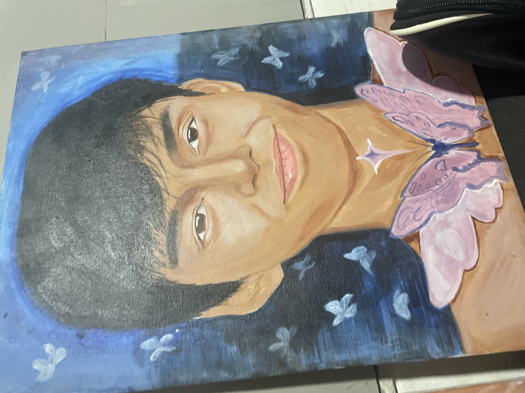 A portrait painting is depicted, showing a young male subject with short, dark hair and medium-brown skin, presented in a head-and-shoulder view. The subject has dark eyes and a gentle smile. The background of the painting features a deep blue hue with lighter blue accents, populated by several small, white-blue butterflies. On the subject's upper chest, two large, light pink butterflies are painted, with darker pink detailing, seemingly integrated into the person's form. Above these butterflies, near the base of the neck, a small, four-pointed, star-like shape in light purple is present. The painting itself is a vertical canvas, possibly acrylic or oil, leaning against a light-colored tiled wall with visible grout lines. A dark baseboard or floor is visible at the bottom, and a portion of a black zippered bag rests on this dark surface, partially obscuring the bottom right of the painting. This scene is located in Davao City, Philippines.