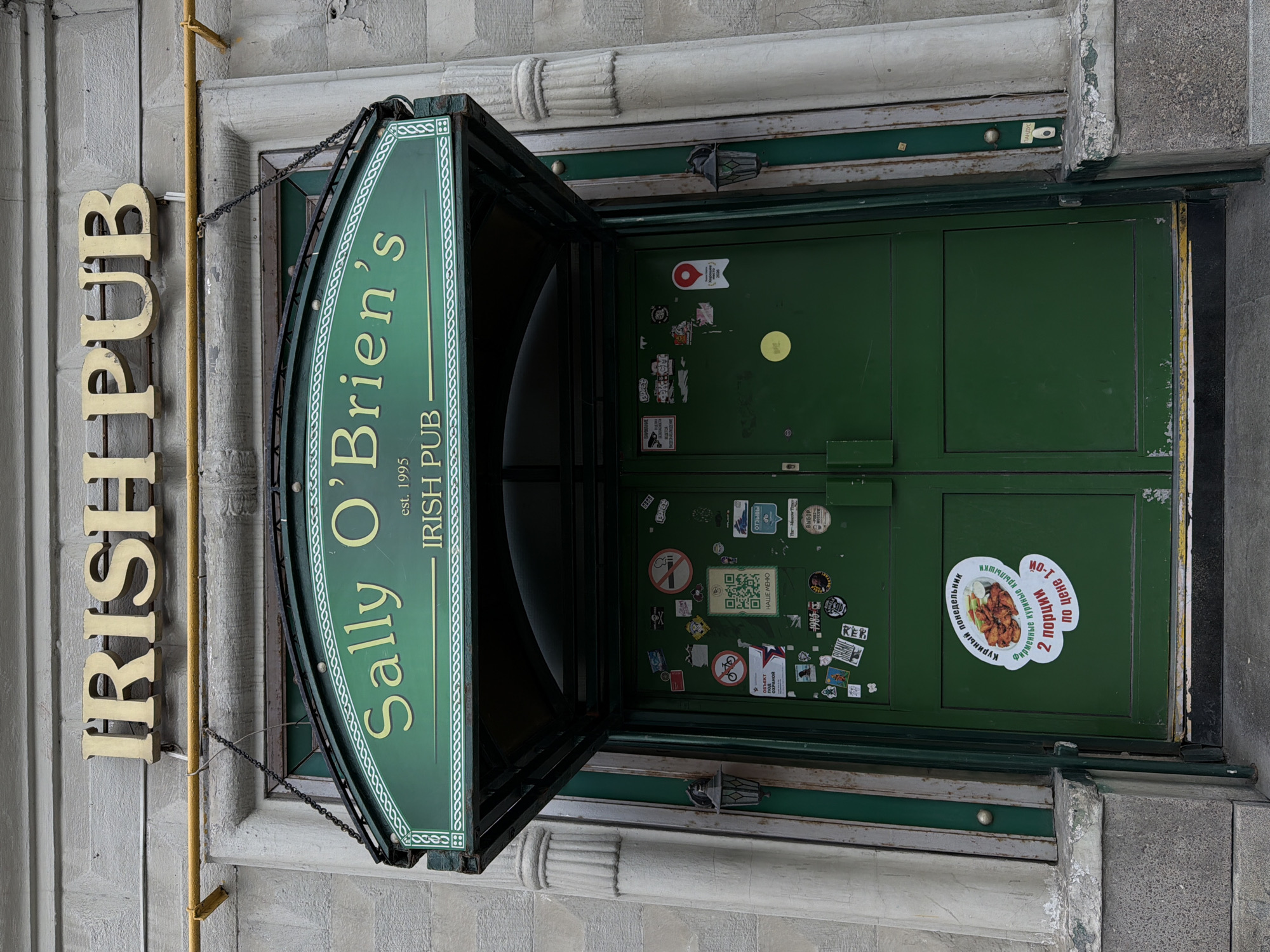 An exterior shot captures the entrance to "Sally O'Brien's Irish Pub" in Moscow, Russia. Above the entrance, a sign reads "IRISH PUB" in large, gold-colored, capital letters. Below this, a green awning with white trim bears the pub's name, "Sally O'Brien's," and "IRISH PUB est. 1995." The double doors of the pub are painted green and are covered with various stickers, including a QR code, a no-smoking sign, a bicycle icon, and what appears to be an advertisement for chicken wings in Russian. Two black, ornate lanterns are affixed to the wall on either side of the entrance. The building's facade is constructed of light-colored stone with some visible wear and tear. The image is taken from a slightly low angle, looking up at the entrance.