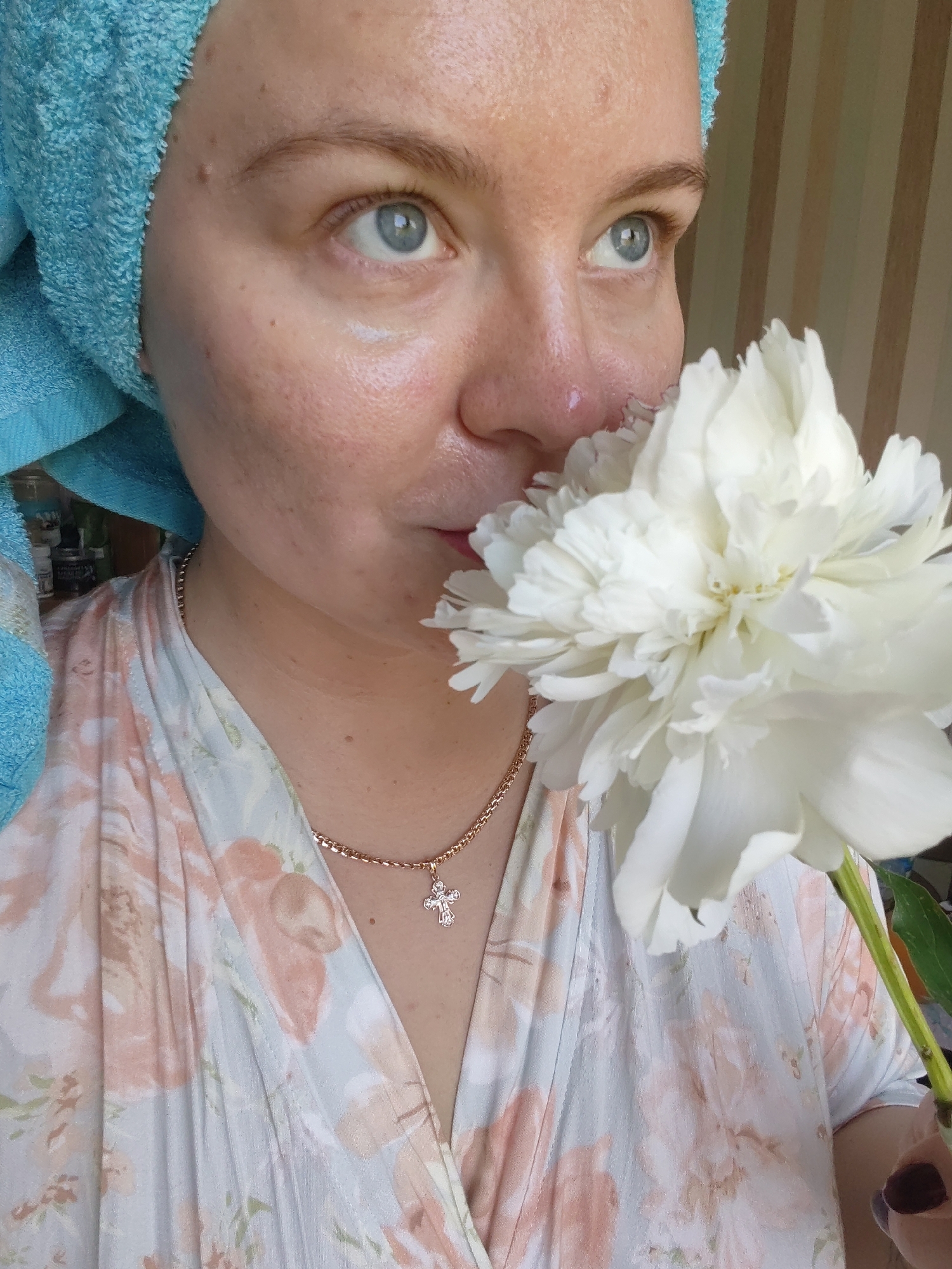 In Izhevsk, Russia, a close-up shot captures a woman wearing a blue towel on her head and a floral-patterned robe. She is smelling a large white peony, holding it near her face. The woman has light blue eyes and is wearing a delicate gold necklace with a cross pendant. The background features vertical stripes, suggesting an indoor setting.