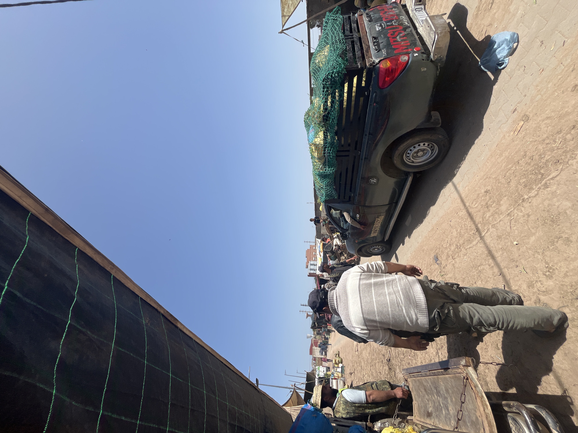 This media file captures a sunny, bustling outdoor market or street scene in Jamaat Shaim, Morocco. The image, originally rotated, shows a clear blue sky dominating the upper left, indicative of midday or early afternoon with strong shadows.
In the foreground, a man is visible from behind, wearing a white and grey striped long-sleeved sweater and olive green cargo pants. He stands next to a rustic wooden cart with metal chains. Another individual, wearing a straw hat and camouflage-patterned clothing, is partially visible near the cart, appearing to be seated or crouched.
To the right, a black Mitsubishi L200 pickup truck is prominently parked. Its bed is heavily loaded with bulky items, likely produce or merchandise, secured under a vibrant green cargo net. The word "MITSUBISHI" and "L200" are clearly visible on the tailgate, along with some red graffiti-like text. The truck has visible wear and tear.
On the far left of the image, a large, dark structure, possibly a stall or a shaded area, is covered with black mesh fabric featuring green stitching, extending vertically. The ground beneath the people and vehicles is a mix of packed dirt and uneven, light-colored paving. In the mid-ground and background, more people, market stalls, and vehicles can be seen, suggesting a vibrant commercial activity. The overall impression is that of a busy, sun-drenched local marketplace.