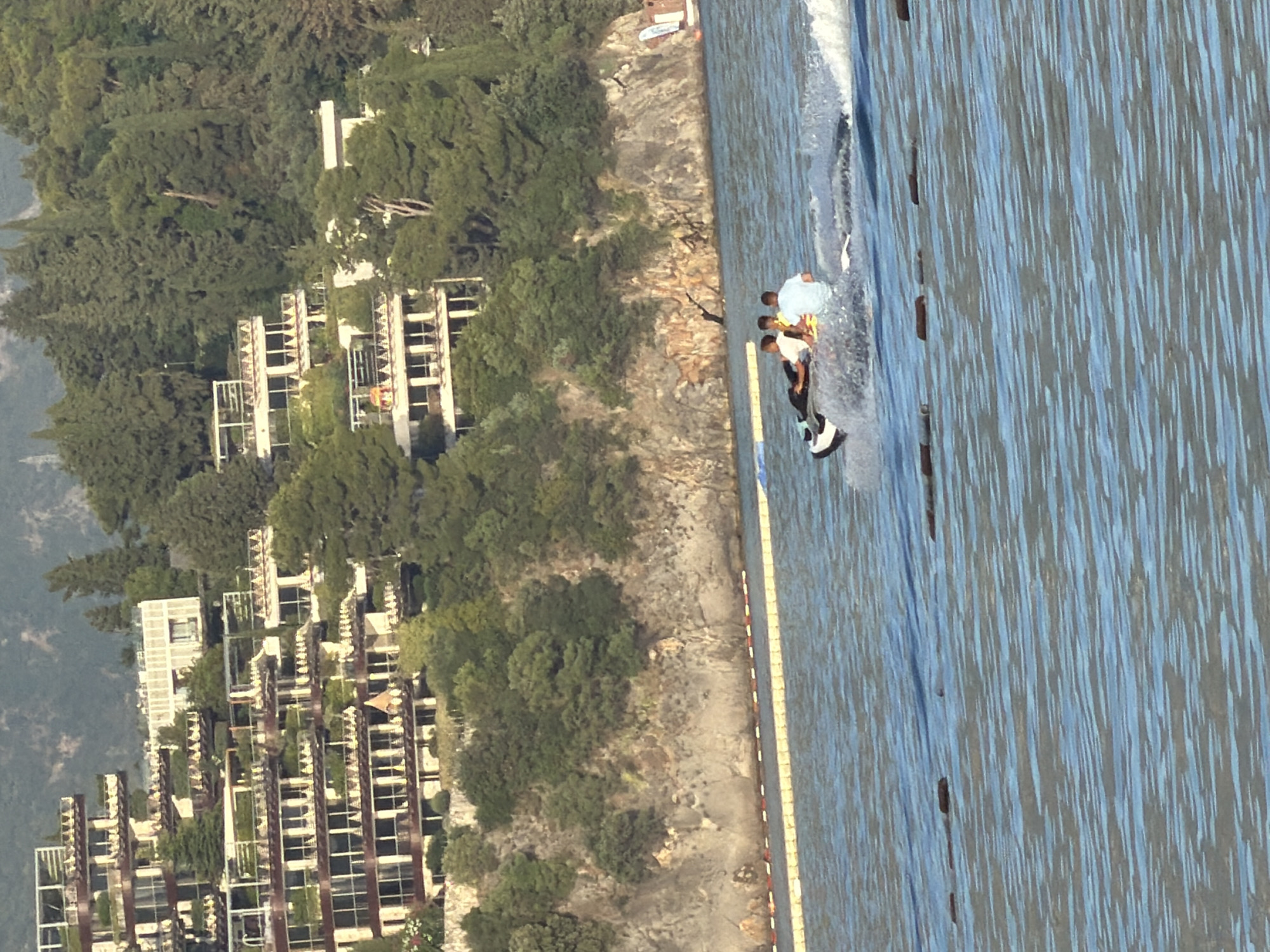 The image, rotated 90 degrees counter-clockwise, depicts a vibrant coastal scene in Budva, Montenegro. On the right, a calm body of blue water, likely the sea, shows ripples and a distinct white wake from a water sport activity. Several individuals are engaged in what appears to be wakeboarding or a similar activity. One person, wearing a light blue shirt, is positioned prominently on a dark and white board, generating the wake. Another person in an orange shirt is situated behind them, and a third person is visible further back. A yellow floating barrier runs parallel to the shore, segmenting the water.
To the left, a steep hillside is densely covered with lush green trees. Numerous multi-story buildings are nestled into this verdant landscape, featuring balconies and a modern, tiered architectural style, suggesting a resort or residential complex. The immediate shoreline between the water and the vegetated hill appears rocky and undeveloped in places. The scene is bathed in bright sunlight, indicating a clear, sunny day, likely morning or afternoon. The overall atmosphere is recreational and leisurely. No text is visible in the image.