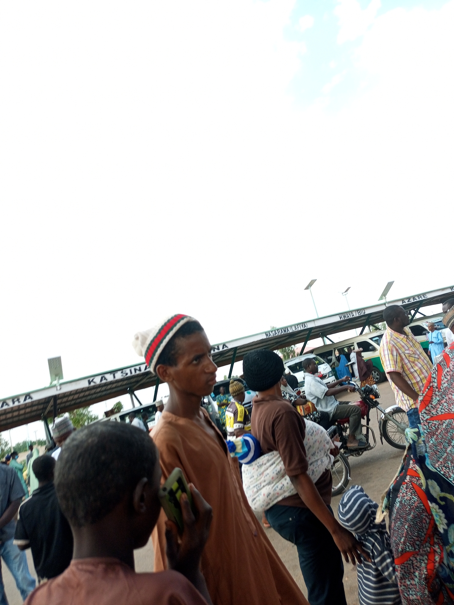 The image depicts an outdoor scene at what appears to be a transportation hub or market in an African city, with a bright, overcast sky. In the foreground, a young boy with short, dark hair holds a green cell phone in his right hand and is looking to the right. He wears a brown tunic. Behind him, a person with dark skin and short hair, wearing a striped beanie and a brown robe, faces right. This person is carrying a baby wrapped in a patterned cloth on their back. To the right of this individual, another person in a colorful patterned dress walks away from the camera.
In the middle ground, several vehicles and people are visible. A man in a yellow and white plaid shirt rides a motorcycle, looking forward. Another man in a light blue shirt stands near a white van. Signs are visible above a covered walkway, with text including "KATSINA," "NASARAWA (TAFIYA)," and "AZARE." Some of these signs are partially obscured. People are seen boarding or disembarking from vehicles. The environment appears to be a paved area with a covered structure, possibly a bus station or a market stall area. The overall impression is one of a busy public space.