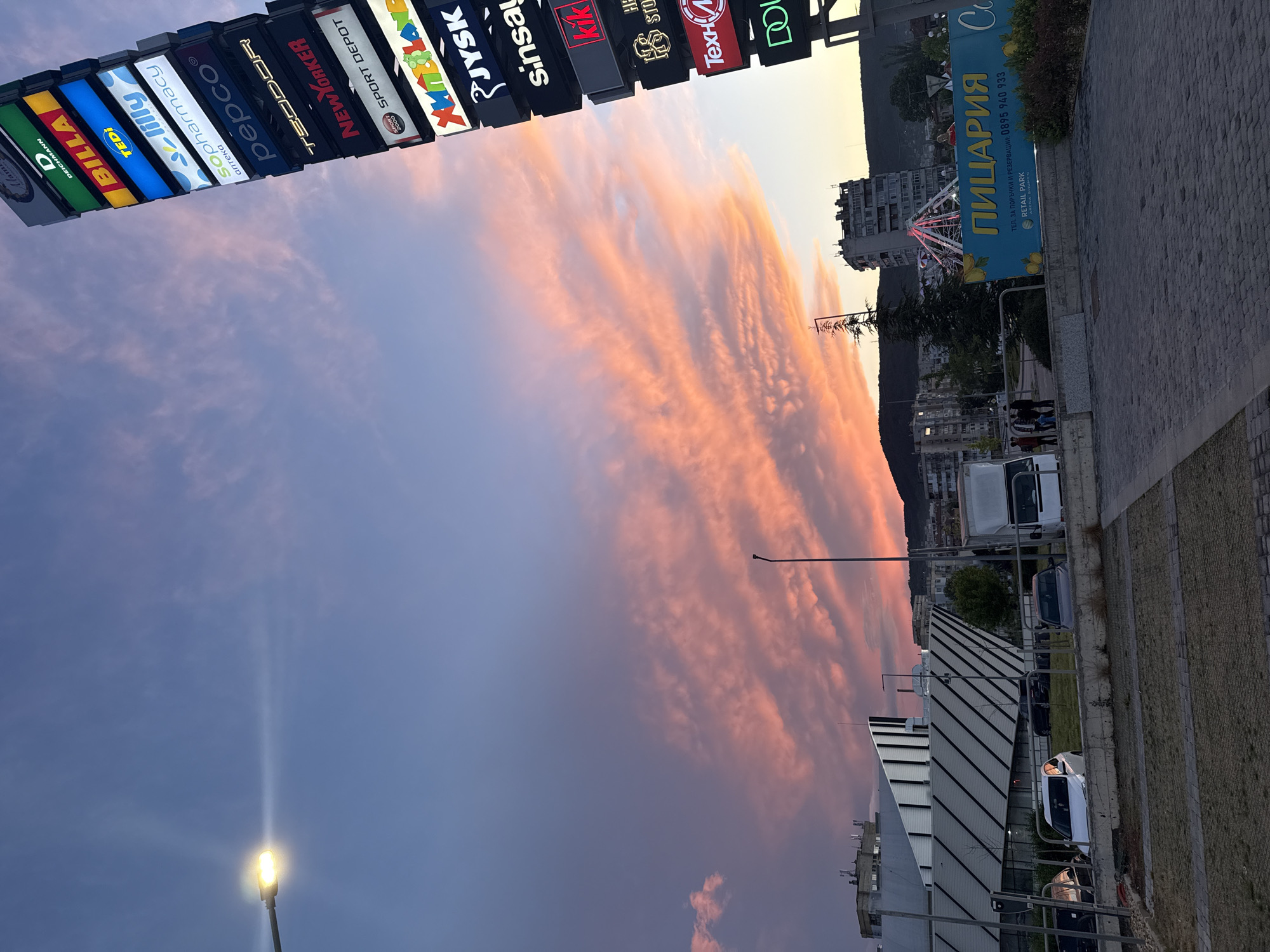 This media file captures an urban commercial area at dusk, featuring a vibrant sunset sky and prominent retail signage. The upper sky transitions from a deep blue on the left to an array of warm oranges and pinks on the right, with elongated, fiery clouds dominating the horizon.
A tall, multi-sided structure along the top displays a vertical stack of illuminated signs for various stores. Visible brands include "DEICHMANN", "BILLA", "TEDi", "Lilly sopharmacy", "pepco", "TEODOR", "NEWYORKER", "SPORT DEPOT", "JYSK", "sinsay", "kik", and "Технополис" (Техни).
In the mid-ground on the right, a large blue billboard advertises "ПИЦАРИЯ" (Pizzeria) with contact information: "ТЕЛ. ЗА ПОРЪЧКИ И РЕЗЕРВАЦИИ: 0895 940 933". Below this, it states "RETAIL PARK ARENA SHUMEN," indicating the location as a retail park in Shumen, Bulgaria. Behind this sign, a brightly lit Ferris wheel is visible among distant buildings and trees.
The lower portion of the image shows architectural structures, including a building with a corrugated roof, and a paved area or parking lot with several vehicles. A few indistinct figures appear in the distance. A streetlamp in the bottom left emits a strong, bright light, casting a visible ray upwards into the blue sky. The scene conveys the busy atmosphere of a retail hub during the beautiful transition from day to night.
