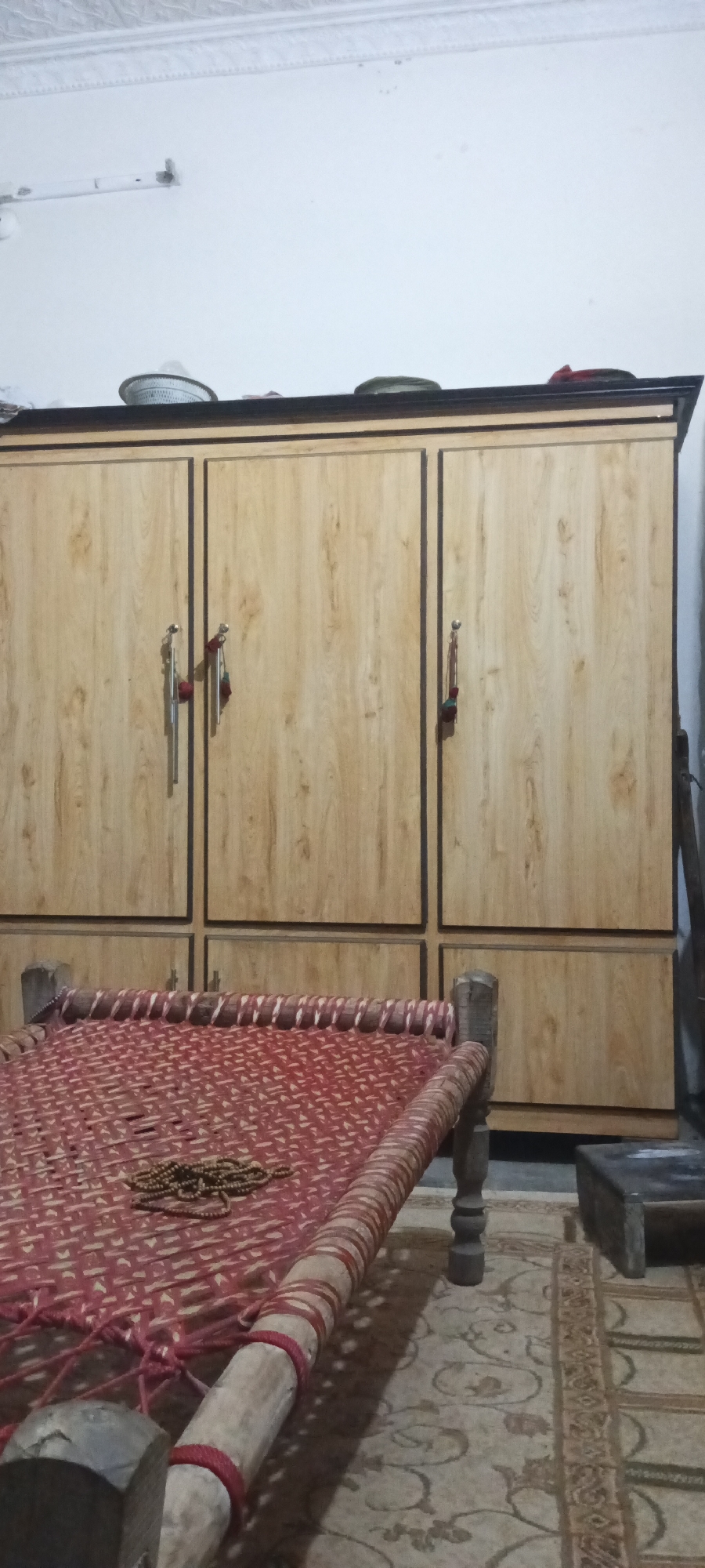 The interior shot depicts a bedroom in Katlang Tehsil, Pakistan. Dominating the scene is a large wardrobe, featuring a light wood grain finish with dark brown accents around the edges and on the handles. On top of the wardrobe are several plates and other items.
In the foreground is a traditional woven bed (charpai) made of light-colored wood with a red and off-white woven surface. A set of prayer beads rests on the bed's surface. The floor is covered with a patterned rug in shades of brown and beige. A dark wooden object is visible on the floor near the wardrobe. The walls are painted white.