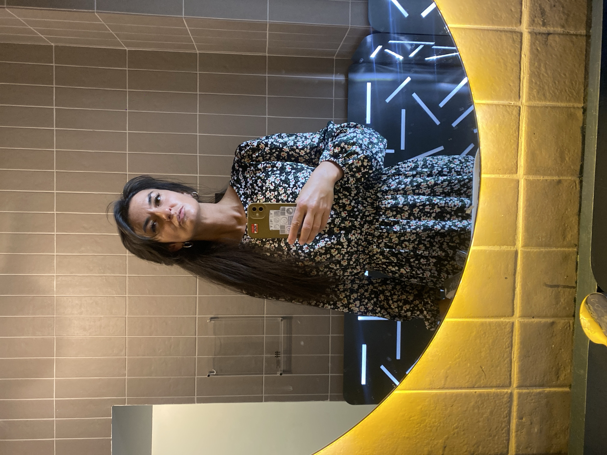 A woman is taking a mirror selfie in what appears to be a restroom. She has long, dark hair and is wearing a long-sleeved dress with a small floral pattern. The phone she's using is gold-colored. The mirror has a distinctive design with a yellow-tiled frame and internal LED lighting. The background includes light-brown, rectangular tiles on the wall, and a design element of illuminated, geometric shapes within the mirror's reflection. While the exact location within Moscow, Russia, is not possible to determine with the given visual information, the overall impression suggests a modern, stylish interior, possibly in a restaurant, hotel, or public space.