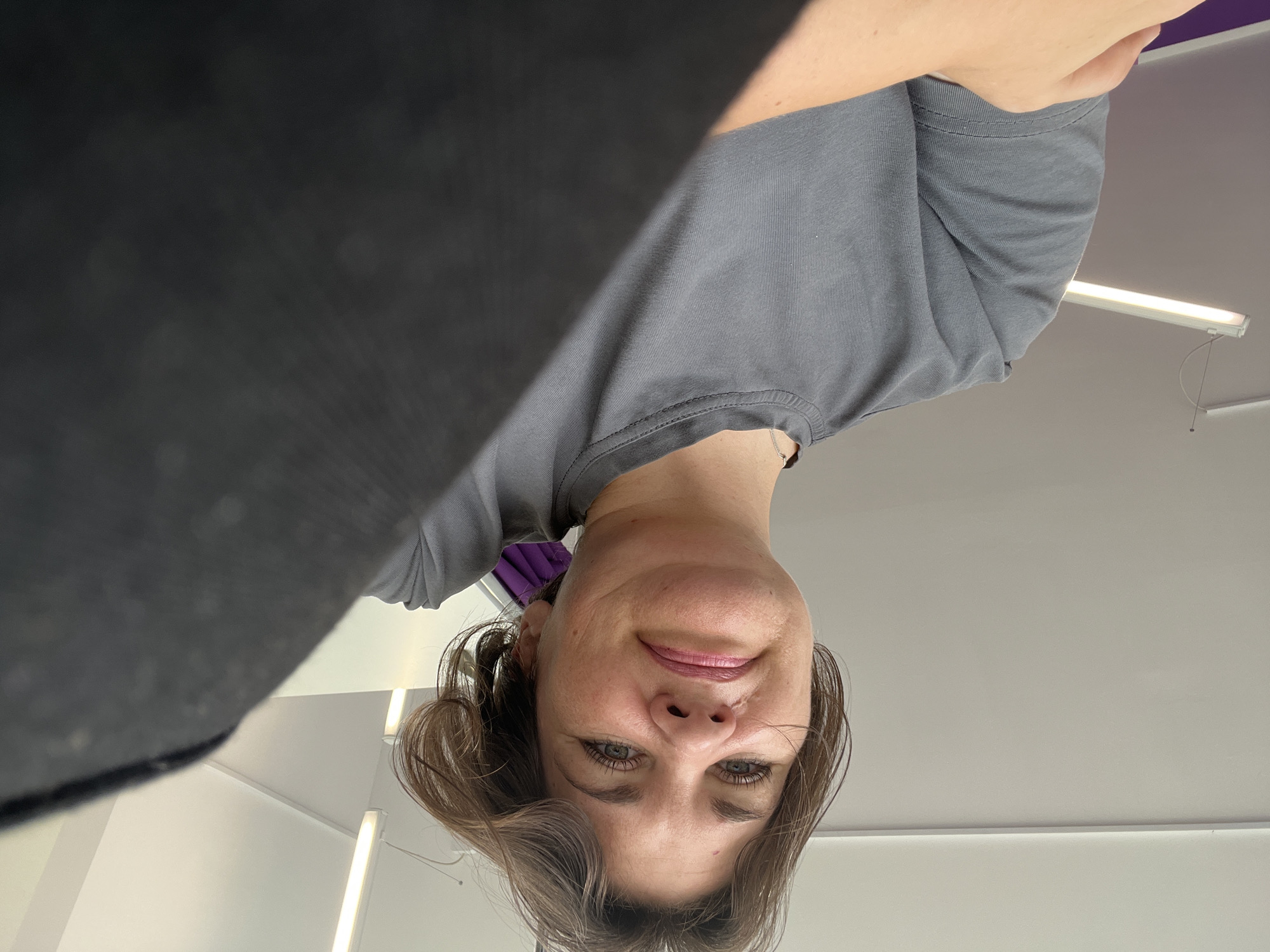 A low-angle, medium shot captures a woman from the chest up. She has light brown, shoulder-length hair that is slightly tousled, and her face is turned slightly upwards towards the camera. Her expression appears neutral to slightly displeased. She is wearing a dark grey t-shirt. The background consists of a white ceiling with fluorescent light fixtures. The image appears to be taken indoors, possibly in a commercial or office space, but no specific location in Afipskiy, Russia is identifiable from the visual information.