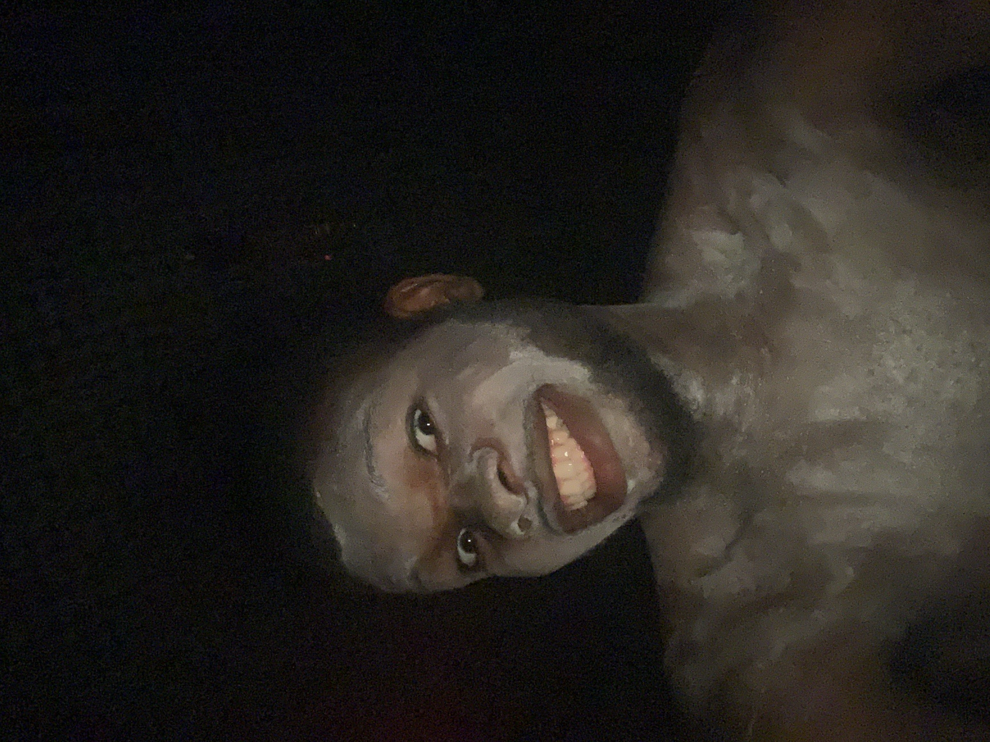 A person with dark skin is smiling, with white paint smeared on their face and upper body. The background is dark. The image does not contain enough information to reliably place this person in Onitsha, Nigeria.