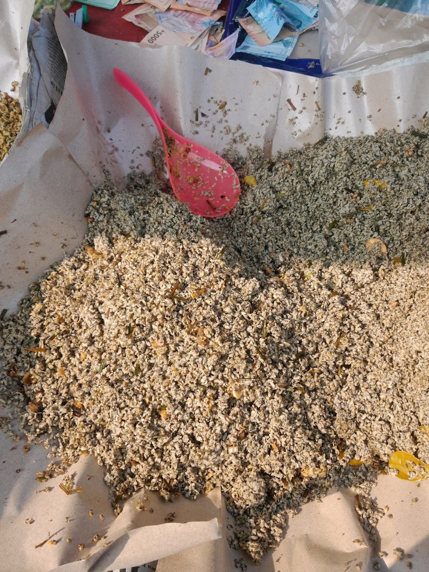 In Jogosatru, Indonesia, a close-up shot reveals a pile of dried herbs and seeds, possibly medicinal or culinary, spread out on brown paper. A vibrant pink plastic scoop rests on the material, suggesting it's being measured, prepared, or dispensed. In the background, a stack of Indonesian currency, including denominations of 5000 and 2000, is visible, indicating a transaction or sale is in progress. The overall scene suggests a marketplace or a vendor's stall where these goods are being sold.