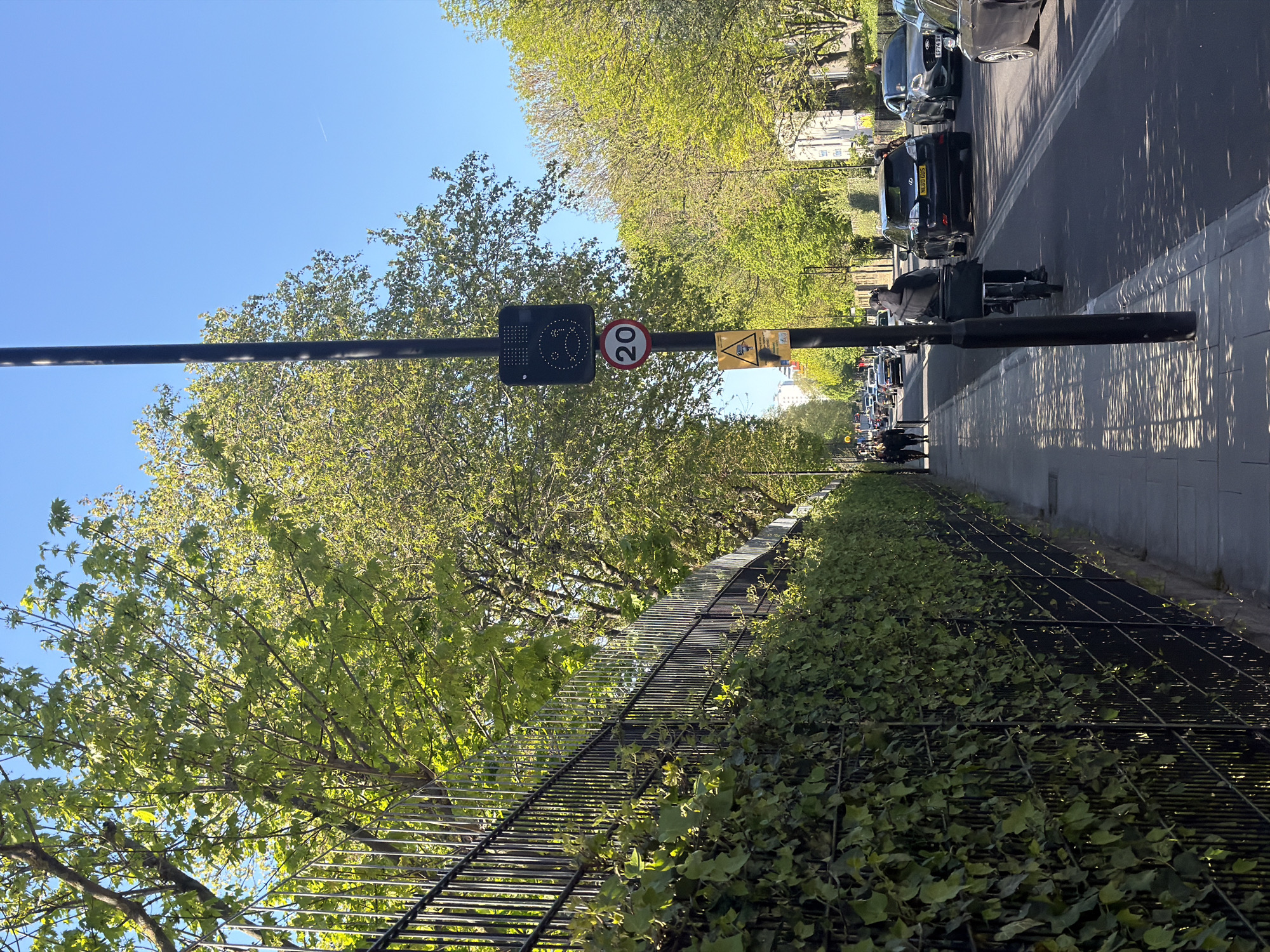 This media file captures a street scene in London, United Kingdom, under bright, clear conditions, likely during spring or early summer given the lush green foliage. The image is oriented as if looking down a tree-lined avenue.
In the foreground, a prominent black pole stands on the right side of the sidewalk, from which a horizontal bar extends across the middle of the frame. Attached to this bar are several signs: an electronic display showing a pixelated graphic (possibly a speed awareness symbol or smiley face), a red-bordered circular "20" mph speed limit sign, and a yellow triangular warning sign with unreadable text.
To the left of the pole, a black metal fence, heavily overgrown with bright green ivy, runs parallel to the street. Beyond this fence and lining the street are numerous mature trees with vibrant green leaves, casting dappled shadows. The sky above is a clear, brilliant blue, with a faint airplane contrail visible in the upper right.
On the right side of the pole, a paved sidewalk transitions to an asphalt road. Several vehicles are present: a black SUV with the license plate "SF13 OZS" is visible, followed by another dark car with a partially obscured license plate "EF74 BV". A person on a bicycle, equipped with a large black delivery box, is positioned near the pole, suggesting a delivery service. Further down the road, additional cars and pedestrians can be seen, indicating a moderately busy urban setting.
The strong sunlight creates distinct shadows from the trees and the horizontal bar across the road and sidewalk. Visible text includes: "20" on the speed limit sign, and the license plates "SF13 OZS" and "EF74 BV" on the vehicles.