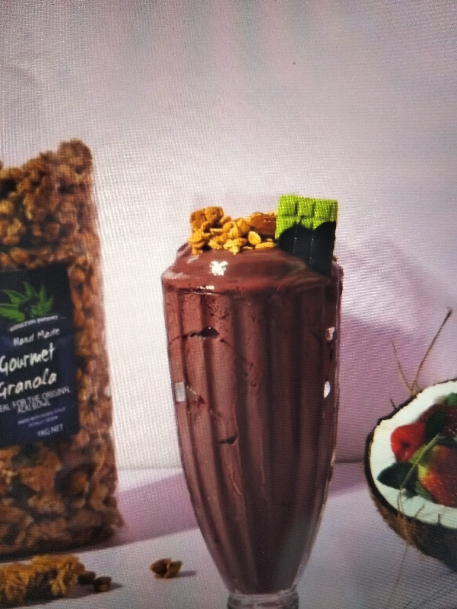 This image depicts a close-up, still-life arrangement of food items on a light-colored surface, against a softly lit, out-of-focus background. In the foreground left, a large, clear plastic bag is filled with granola, featuring a dark blue label with white text and a green leaf logo. To the right of the granola bag is a tall, fluted glass filled with a thick, dark brown liquid, likely a milkshake or smoothie. The surface of the drink is topped with a small pile of granola clusters and a piece of green-covered chocolate. Further to the right, a halved coconut is visible, revealing fresh strawberries and some greenery inside. Scattered granola pieces are present on the surface in front of the glass. The overall impression is a product display, emphasizing healthy and indulgent food options. There is no indication of a specific city, including Payagaboh, Indonesia, or any people present in the scene.