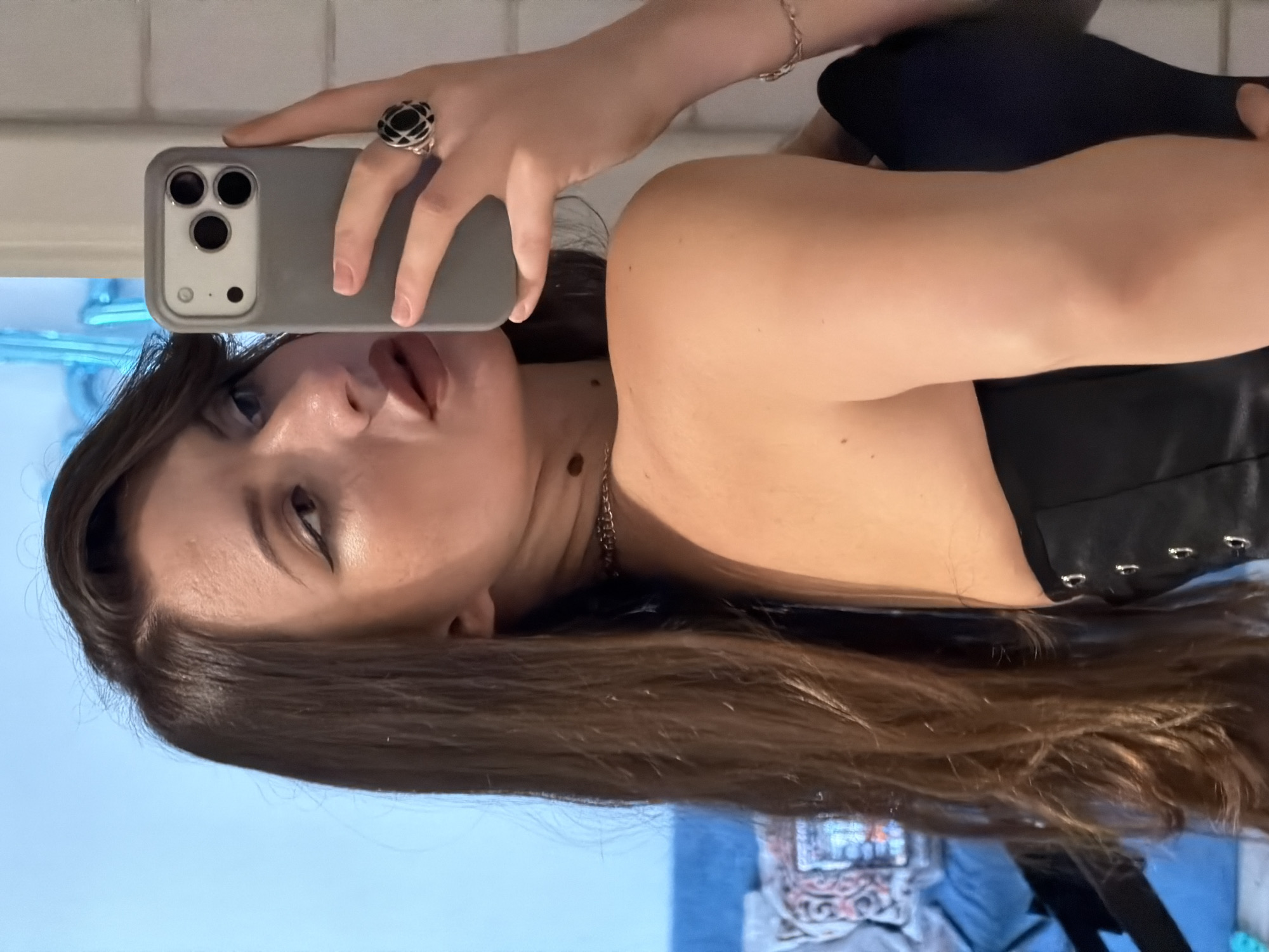 A young woman with long, dark brown hair and dark-winged eyeliner takes a selfie in a well-lit indoor space. She is wearing a black corset and a silver chain necklace. Her right hand is holding a grey smartphone, and she is wearing a large, ornate black and silver ring on her index finger. A silver bracelet is visible on her left wrist. The background shows a blue wall, a doorway, and what appears to be a blurred couch with a patterned throw pillow. The image does not contain any visual cues that would allow for the determination of its location within Sharm El Sheikh, Egypt.
