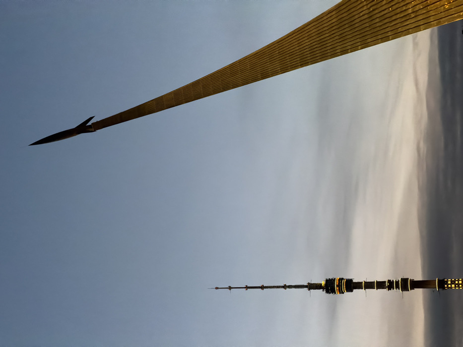 The image displays two prominent landmarks in Moscow, Russia, near the VDNKh exhibition complex, under a twilight or dawn sky. On the left, the Ostankino Tower, a tall, dark, slender communication tower, rises vertically, featuring segmented sections illuminated with yellow-orange light. On the right, the Monument to the Conquerors of Space, a golden, ribbed structure, ascends diagonally from the lower right to the upper left, culminating in a dark, pointed rocket at its peak. The sky transitions from a light, clear blue at the top to a darker, cloud-strewn grey-orange closer to the horizon. No people, specific actions, or interactions are visible in the scene.