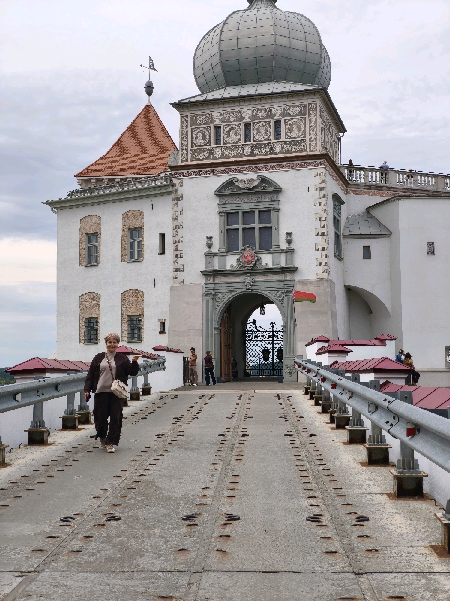 This image depicts the entrance to a castle with a woman walking across a bridge towards it. The castle is a large white structure with a distinctive domed tower and a red-tiled roof. There are several windows and architectural details visible on the castle's facade. The bridge is made of metal and concrete, with railings on either side. Other people are visible in the background, some standing near the castle entrance and others on a balcony above. A flag, appearing to be the flag of Belarus, is attached to the castle wall. The sky is overcast. The location is likely the Kosava Castle, located in the Ivatsevichy District of Brest Region, Belarus, as the architecture and flag are consistent with this landmark.