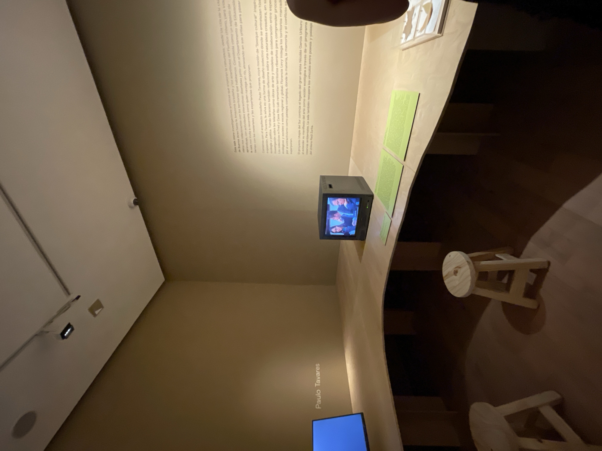 The image depicts an art exhibition in Buenos Aires, Argentina featuring the work of Paulo Tavares and other artists. The location is an indoor space with a high ceiling and beige-colored walls. The exhibition features a curved wooden structure that functions as a display surface. This structure holds a small CRT television displaying a video and several green panels with text.
In the foreground, there are two small wooden stools. The scene is well-lit, and the composition is from a high angle. The text on the wall provides context about the exhibition, mentioning the exploration of nature from Buenos Aires to the Brazilian Amazon and the Chilean cliffs. Other artists mentioned include Tomás Saraceno, m7red, Casa Rio Lab, and Florencia Levy.