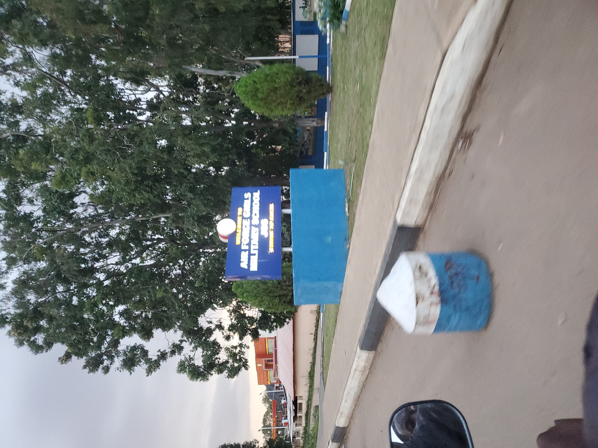 The image shows a street view in Gura Top, Nigeria, featuring a sign for "AIR FORCE GIRLS MILITARY SCHOOL." The sign is blue with white lettering and is mounted on a blue concrete base. In front of the sign, a patch of well-maintained green grass is visible, bordered by a white and blue fence. Trees and other vegetation surround the area. A road with a curb and sidewalk runs in the foreground, with a concrete barrier painted blue and white in the middle of the road. The sky is overcast. In the reflection of a side mirror, a person's face is partially visible. In the background, a building with an orange facade and a red roof is present, along with what appears to be a gas station.