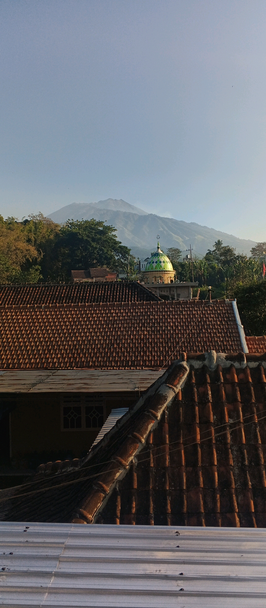 The image depicts a view of Bocek, Indonesia, dominated by a vast, clear blue sky. In the middle ground, a prominent mosque with a green and gold dome stands out amidst lush green trees and residential rooftops. The foreground is filled with the textures of overlapping terracotta roof tiles and a corrugated metal roof. In the distance, a hazy mountain range creates a scenic backdrop, suggesting a location with natural beauty. The scene is illuminated by natural daylight, casting shadows and highlighting the details of the architecture and landscape. There are no people visible in the image.