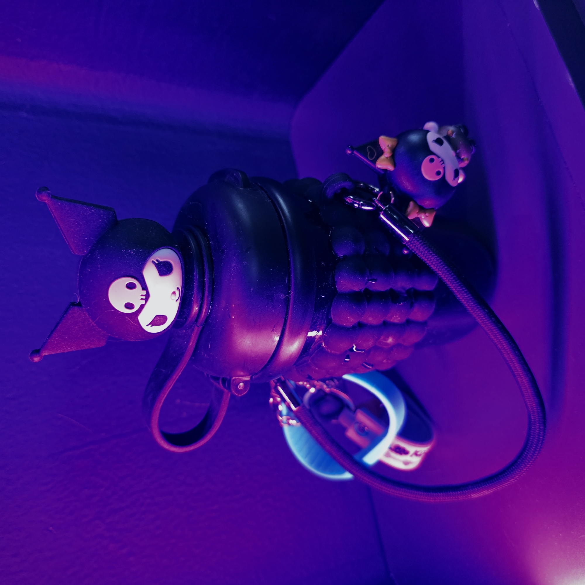 A close-up shot features two stylized black figures against a vivid purple and magenta backdrop. The larger figure, positioned prominently on the left, has pointed ears or horns and a white, skull-like face with large eyes and a wide, grinning mouth, strongly resembling the Sanrio character Kuromi. To its right, a smaller, similarly designed black figure is visible, also with a white skull face, small pointed ears, and adorned with two yellow bows – one on its head and another at its neck. This smaller figure is connected to the larger one by a silver chain.
Between the two figures, and extending to the lower right, is a black, textured object with a pattern of small, rounded protrusions, possibly part of a device or accessory. A braided black cable, reflecting purple light, emerges from near the larger figure and curves downwards, leading towards a bright blue glowing light source in the lower right, which appears to be a ring or strip of light.
The scene is lit by intense, artificial purple and magenta light, creating deep shadows and luminous highlights, particularly on the contours of the objects and the characters' white faces. The background is completely out of focus, showcasing smooth gradients of these vibrant colors, suggesting an indoor, low-light setting with mood lighting. There is no specific activity depicted, and no visible text. While the location context of Saint Petersburg, Russia, is provided, there are no visual cues within the image to confirm this. The overall aesthetic is dramatic, moody, and highlights the "cute but spooky" design of the characters.