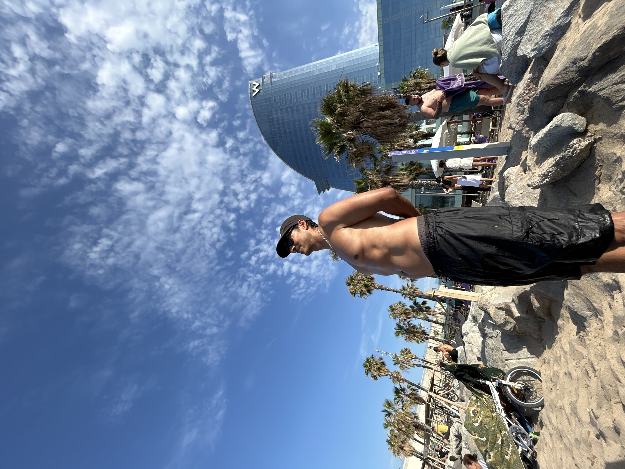 A low-angle, full-shot photograph captures a man standing on a rocky, sandy beach with his back to the camera, facing a bright blue sky filled with scattered white clouds. He is shirtless, wearing black athletic shorts, a dark baseball cap, and sunglasses. His hands are clasped behind his back. In the background, a modern, sail-shaped glass skyscraper with a prominent "W" logo on its facade stands tall. Palm trees are scattered throughout the scene, some in front of the building and others lining the beach. Several other people are visible in the middle ground and background, some sitting on the rocks, others standing or walking near beach umbrellas and loungers. To the left of the main subject, a bicycle is parked on the sand, partially covered by a patterned blanket. The overall scene depicts a sunny day at a beach resort area in Barcelona, Spain, likely near the W Hotel.