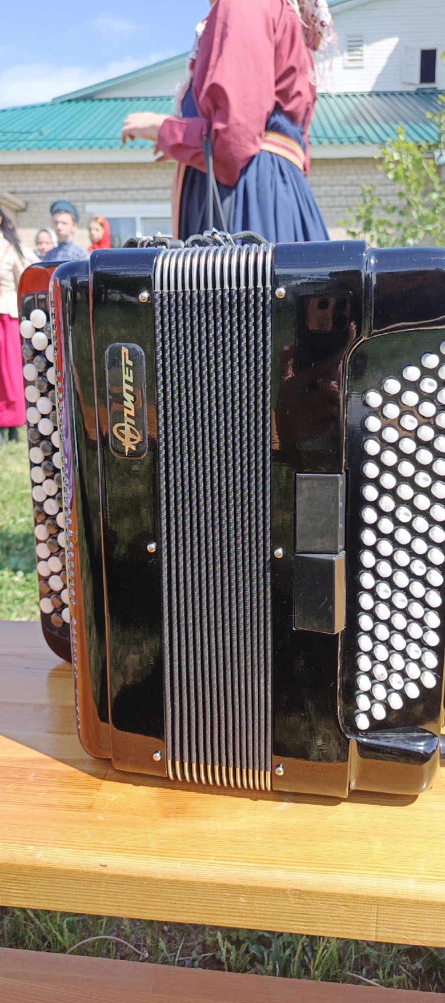 A black and silver "Jupiter" button accordion sits on a wooden surface in an outdoor setting. The accordion's bellows are visible. A person wearing traditional red and blue clothing stands in the background, along with a group of individuals. The location is likely Кемуль, Russia, given the logo on the accordion. The accordion is on a wooden surface. Green grass is visible around the wooden surface.