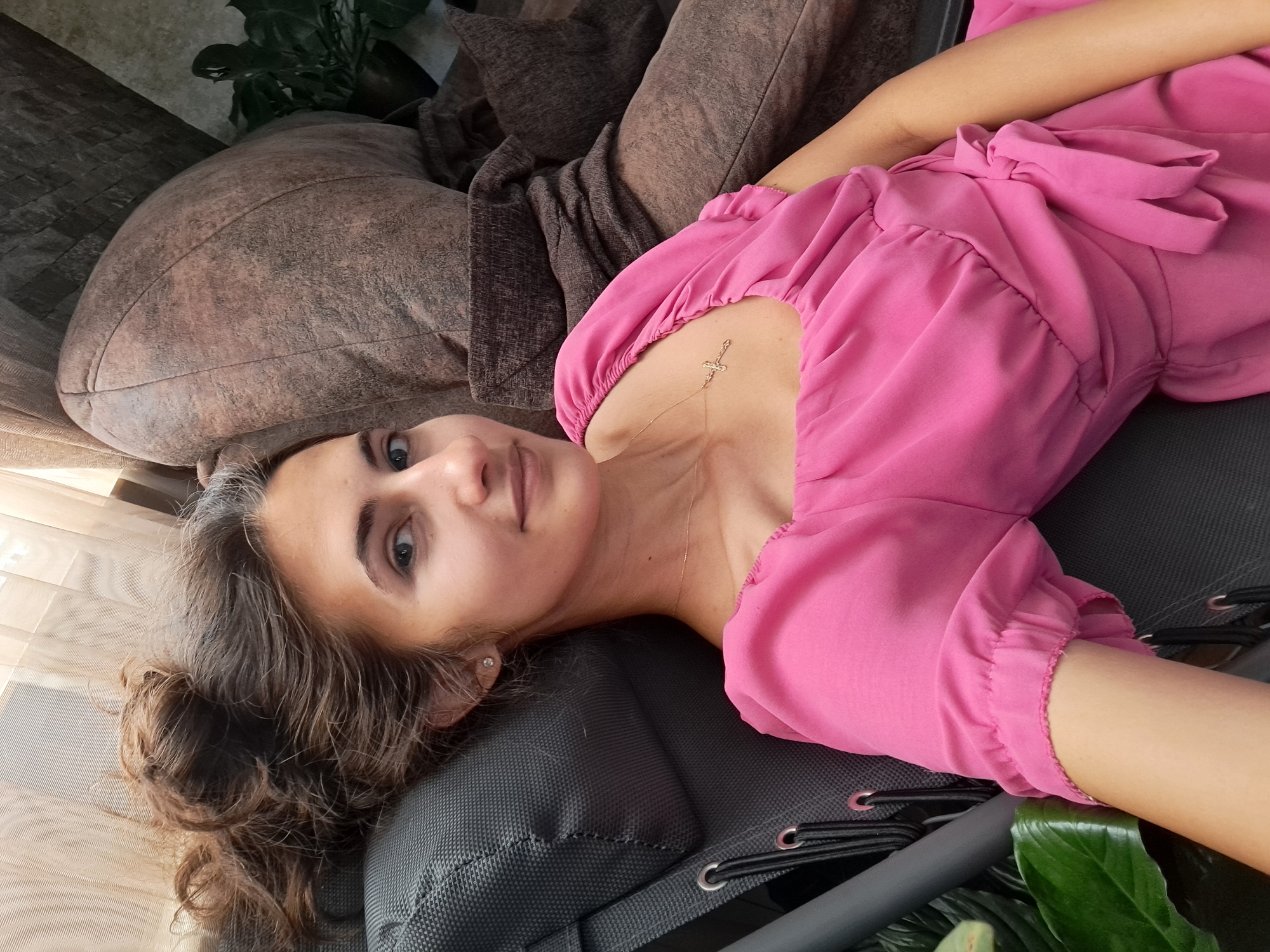 A woman with brown hair pulled up in a messy bun and blue eyes is reclining in a chair. She is wearing a pink dress with ruffled sleeves and a bow at the waist. A gold cross necklace is visible around her neck. The chair appears to be a lounge chair with a gray fabric seat. Behind her, a large brown upholstered piece of furniture, possibly a sofa or armchair, is visible. A plant with large green leaves is in the lower left corner of the image. The background includes sheer curtains and a textured wall.