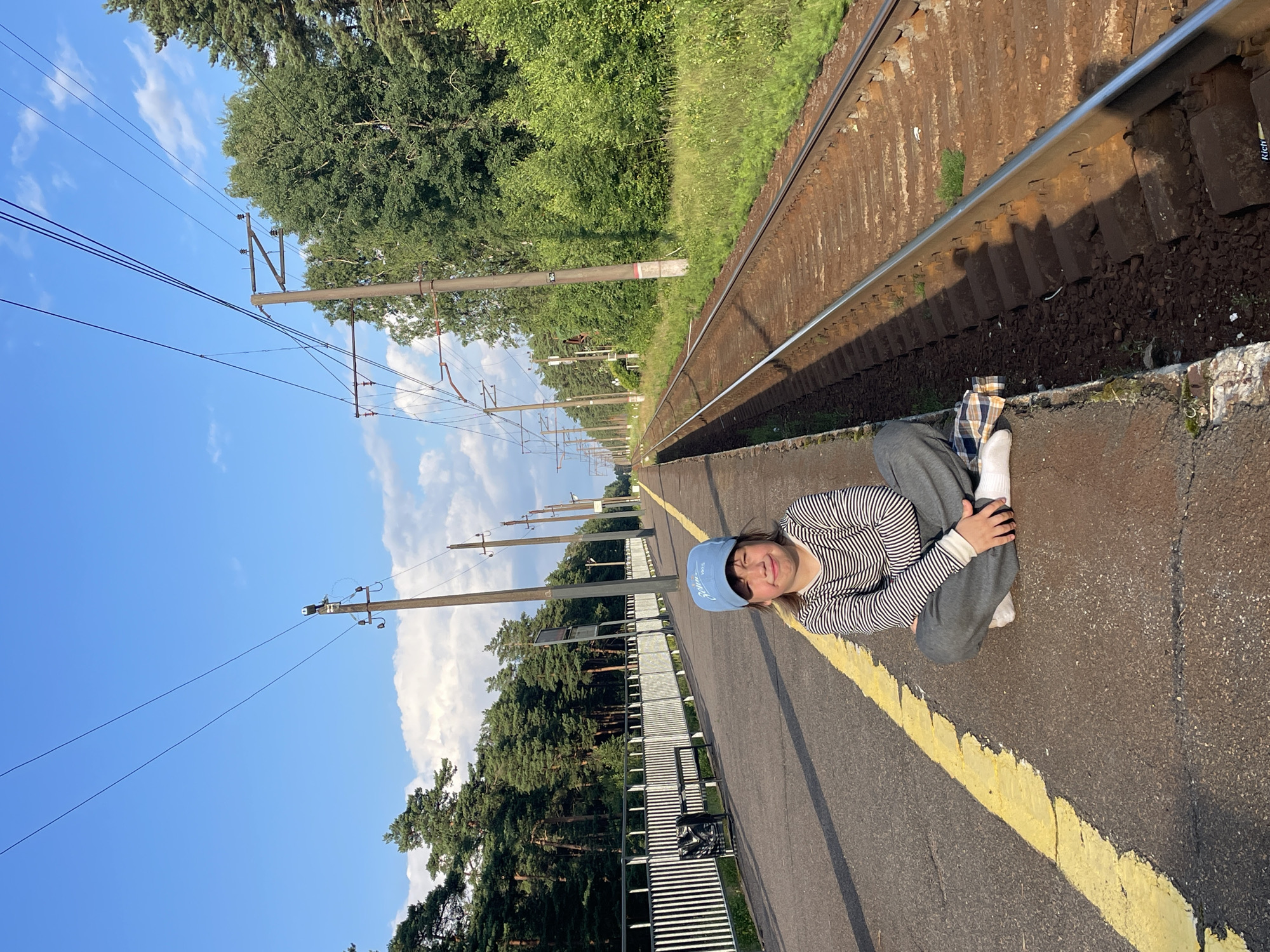 A young person wearing a blue baseball cap and a striped long-sleeved shirt is sitting cross-legged on a train platform in Lebyazhye, Russia. The platform is adjacent to a set of train tracks, with overhead power lines running parallel to the tracks. A white fence is visible behind the person, separating the platform from a dense forest. The sky is clear and blue with a few white clouds. The sun is casting long shadows, indicating it is either early morning or late afternoon. The ground on the platform is asphalt with a yellow line marking the edge. The train tracks are made of ballast and wooden ties.