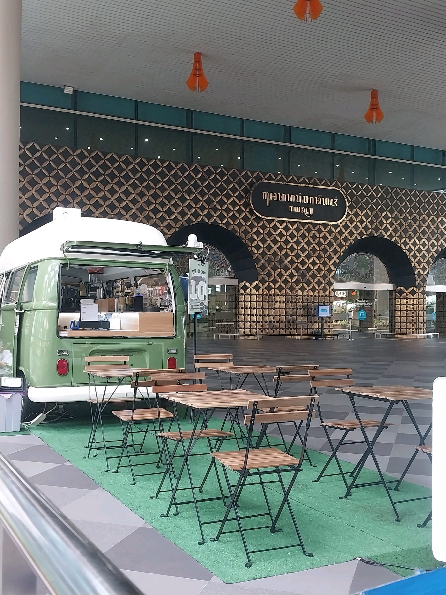 In Palihan, Indonesia, a green vintage Volkswagen van, converted into a mobile coffee shop named "DR. KOPI," is parked outdoors. The van is open at the back, revealing a food service counter with equipment. In front of the van, several folding tables and chairs are set up on a green artificial turf area. The background features a modern building with a distinctive patterned facade and arched entrances. Two bright orange decorative lights hang from the ceiling above the scene. The lighting suggests it is daytime.