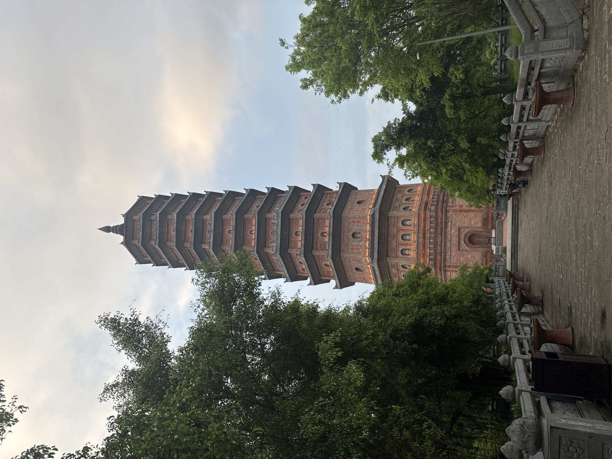 The image depicts the Trấn Quốc Pagoda in the city of Tây Hoa Lư Ward, Vietnam. The pagoda is a towering, multi-tiered structure made of red brick, with intricate architectural details and small illuminated windows on each level. It is surrounded by lush green trees, creating a contrast between the man-made structure and the natural environment. A stone pathway leads up to the pagoda, bordered by a stone railing adorned with lotus-shaped finials. The sky above is overcast with hints of sunlight breaking through, suggesting it may be early morning or late afternoon. No people are visible in the frame, and the scene is peaceful and serene. The overall impression is one of historical significance and architectural grandeur within a tranquil setting.