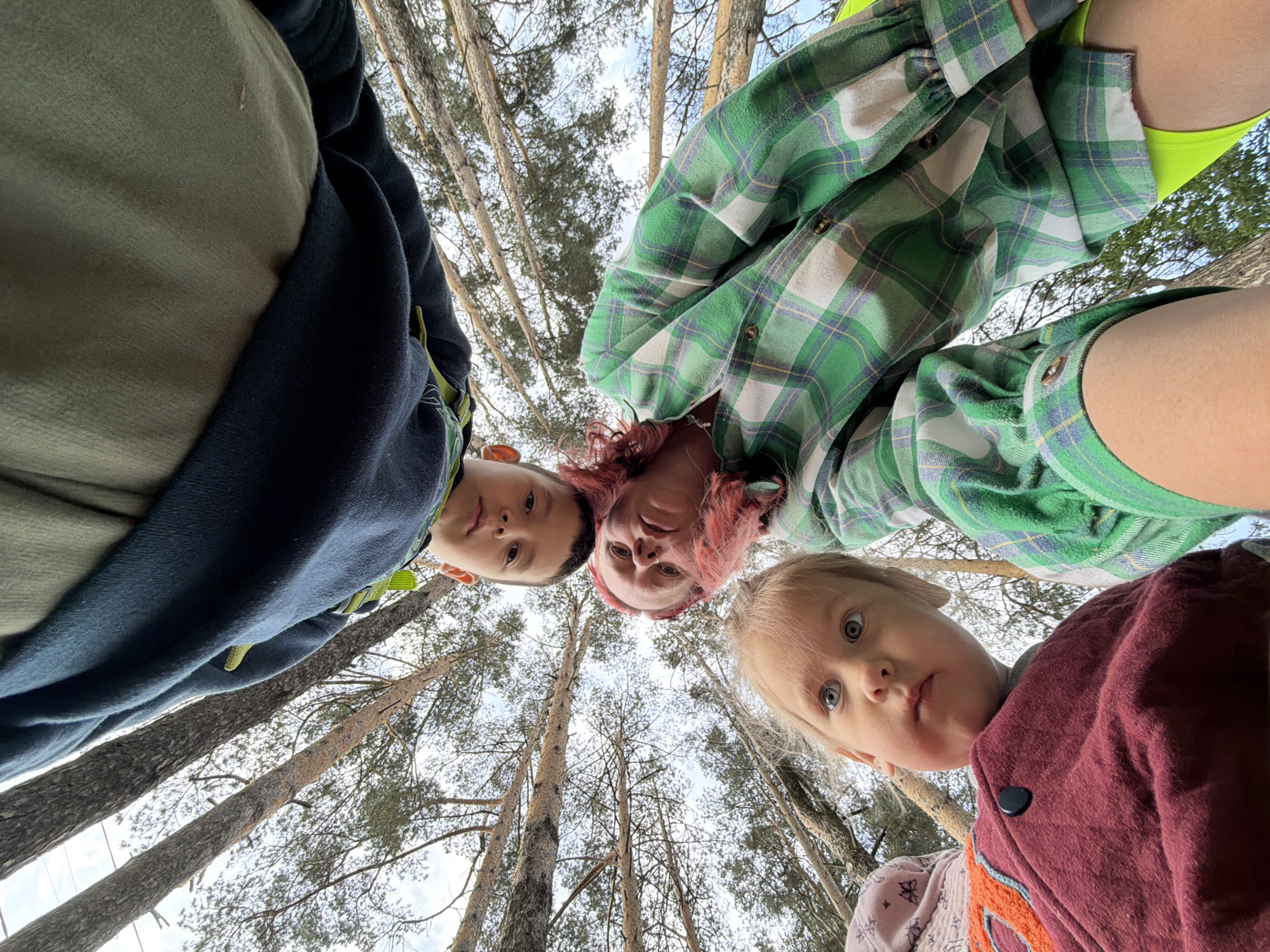 Three individuals, potentially a family, are positioned looking downward towards the camera. The youngest individual is wearing a maroon and orange patterned jacket and has fair hair. Another individual is wearing a green plaid shirt with a neon yellow top underneath and has reddish-pink hair. The third person is wearing a dark blue and green top. The backdrop is filled with tall pine trees. The location is Chaikovsky, Russia.