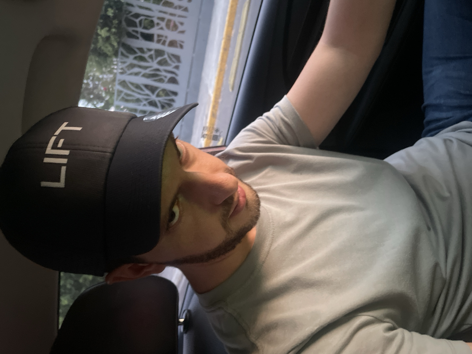 A medium shot of a man with fair skin, wearing a black baseball cap with the word "LIFT" in white block letters, a gray t-shirt, and blue jeans, is seated inside a car. He is facing the camera with a slight tilt of his head. His facial features include a thin beard and mustache. Outside the car window, a glimpse of a white decorative structure and greenery can be seen, suggesting the possible location of Puebla, Mexico. The car's interior is dark, with black seats and a visible door frame.