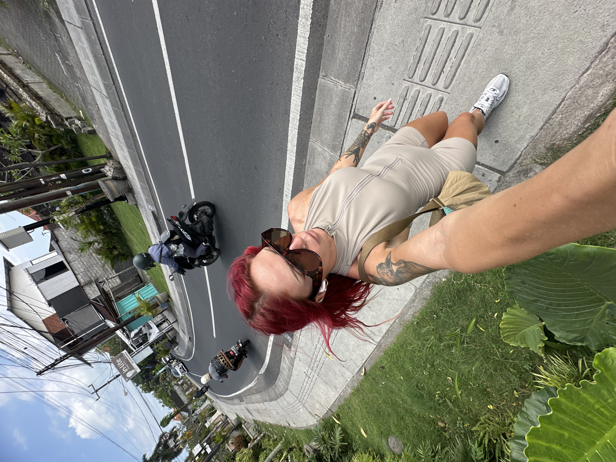 A woman with red hair and sunglasses stands on a sidewalk in North Kuta, Indonesia, taking a selfie. She is wearing a beige jumpsuit, white sneakers, and a tan shoulder bag. Tattoos are visible on her arms. Behind her, a road curves with motorcycles driving on it. A building and utility poles are visible in the background. Grass and large leafy plants surround the sidewalk.