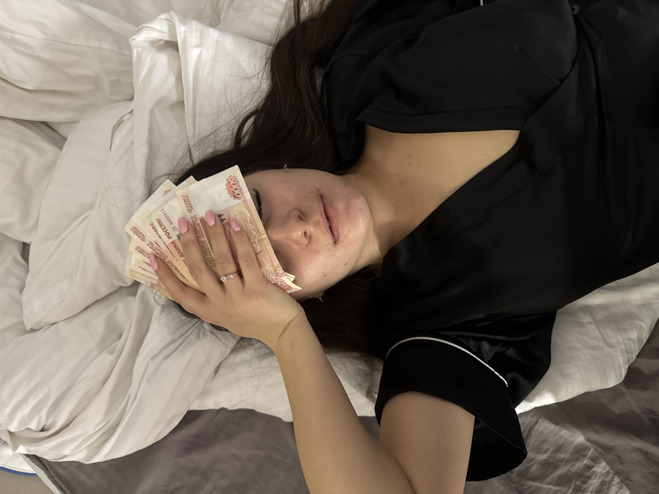 A woman is lying on a bed with a wad of Russian 5000 Ruble banknotes covering one eye. She has light pink nails and is wearing a ring on her ring finger. The woman is wearing a black satin pajama top with white piping. The bedding appears to be mostly white with a section of gray bedding visible in the lower right corner. Her dark hair is spread across the pillow.