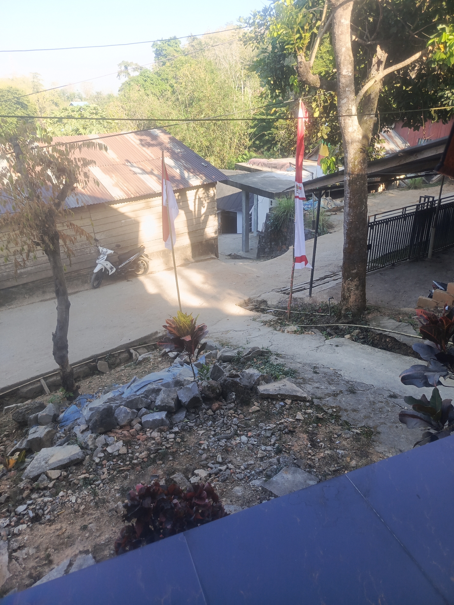 The image depicts an outdoor scene in Kendari, Indonesia, on a sunny day. In the foreground, a rough, gravelly patch of ground is visible, interspersed with larger rocks and some sparse vegetation. A dark blue, flat surface, possibly a railing or a building element, occupies the bottom right portion of the frame.
Further into the scene, a cracked concrete pathway leads towards a cluster of buildings. Two Indonesian flags, one white and one red, are mounted on poles and are positioned near a large tree with a rough, textured trunk. A white motorcycle is parked on the pathway.
The buildings appear to be constructed with wooden walls and corrugated metal roofs. In the background, lush green vegetation covers a hillside, suggesting a natural, possibly semi-rural or suburban environment within Kendari. Power lines are strung across the sky. The lighting indicates it is daytime, with shadows cast by the trees and structures.