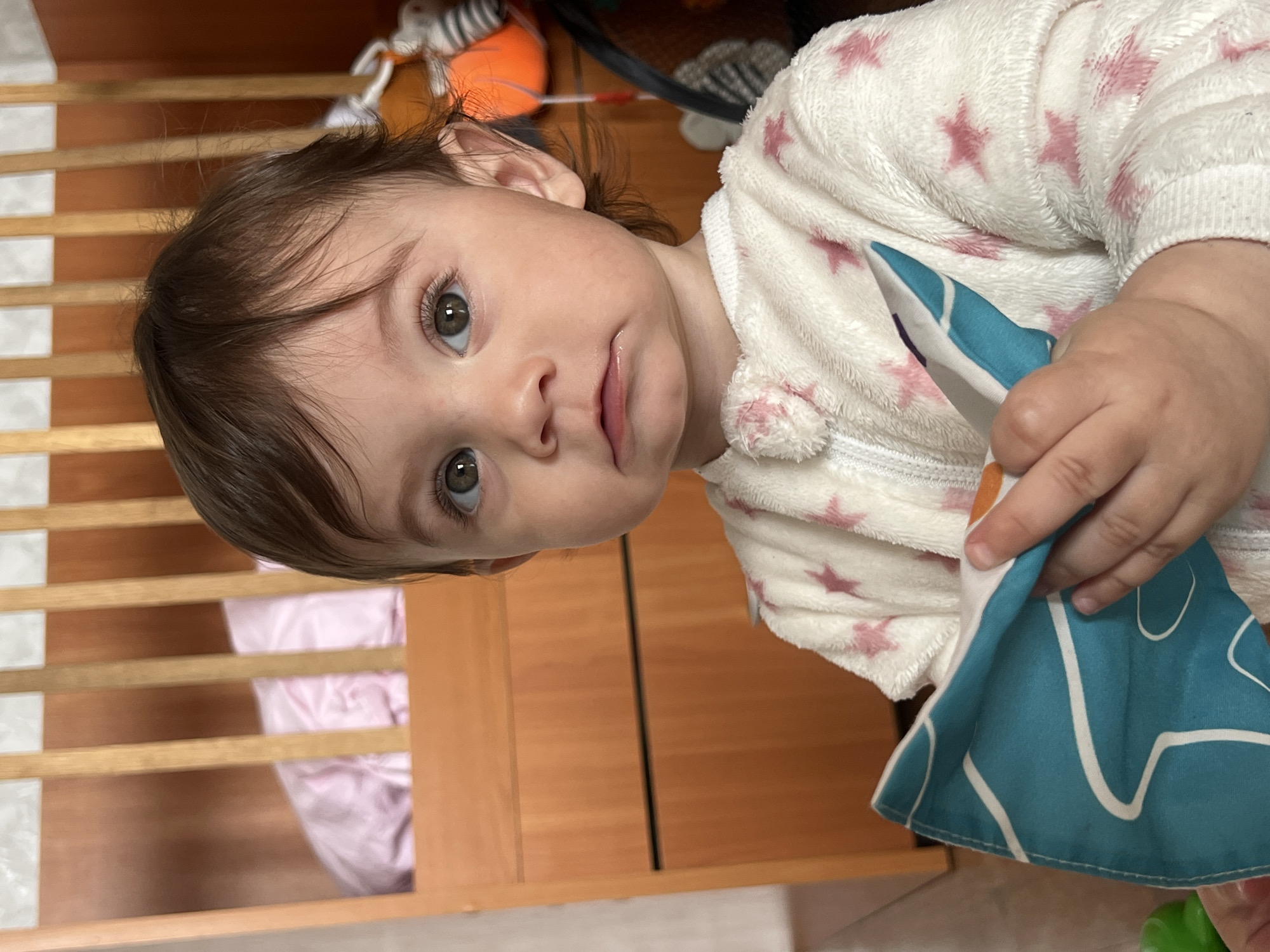 A young child with brown hair and light eyes looks at the camera. The child is wearing a white, star-patterned, zip-up garment and holding a blue and white patterned cloth. A wooden crib is behind the child, and a light wood-colored cabinet or dresser is to the side. It is not possible to verify that this image was taken in Elena, Bulgaria.