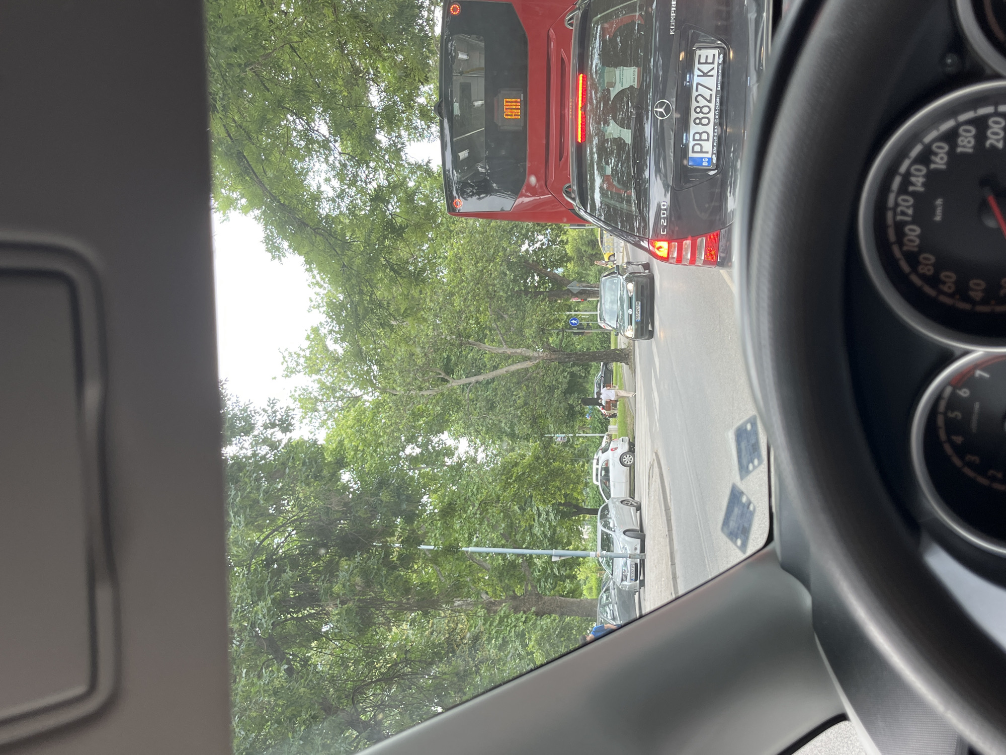 The image captures a traffic scene from the perspective of a car driver in Orizari, Bulgaria. In the foreground, the steering wheel and part of the dashboard are visible. Ahead, a black Mercedes-Benz C200 KOMPRESSOR is in traffic, with its license plate reading "PB 8827 KE". To the left of the Mercedes, a dark green Volkswagen Golf is also in traffic, with its license plate partially visible as "PB 5497 BM". Further ahead, a red bus is stopped in traffic. The surrounding environment features mature trees lining a paved road. Two small square stickers are visible on the interior of the car's windshield. The sky is overcast.