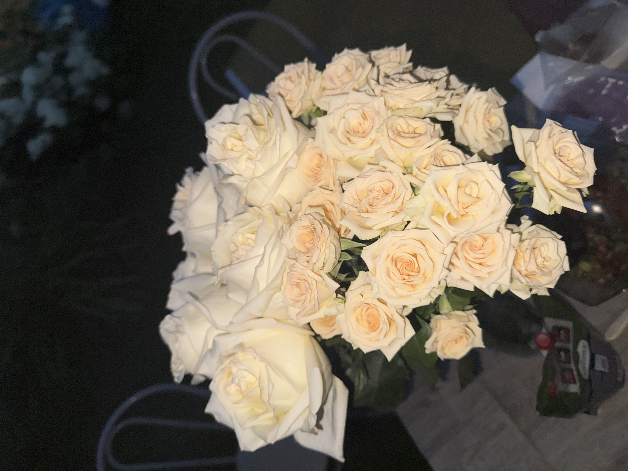 A bouquet of pale cream and peachy roses is arranged in a dark vase on a textured surface, possibly a table or floor. The background is dark, with the blurry shapes of chairs and some foliage suggesting an outdoor or dimly lit indoor setting. The flowers are the primary focus, with some larger roses at the top and smaller roses filling the bouquet.