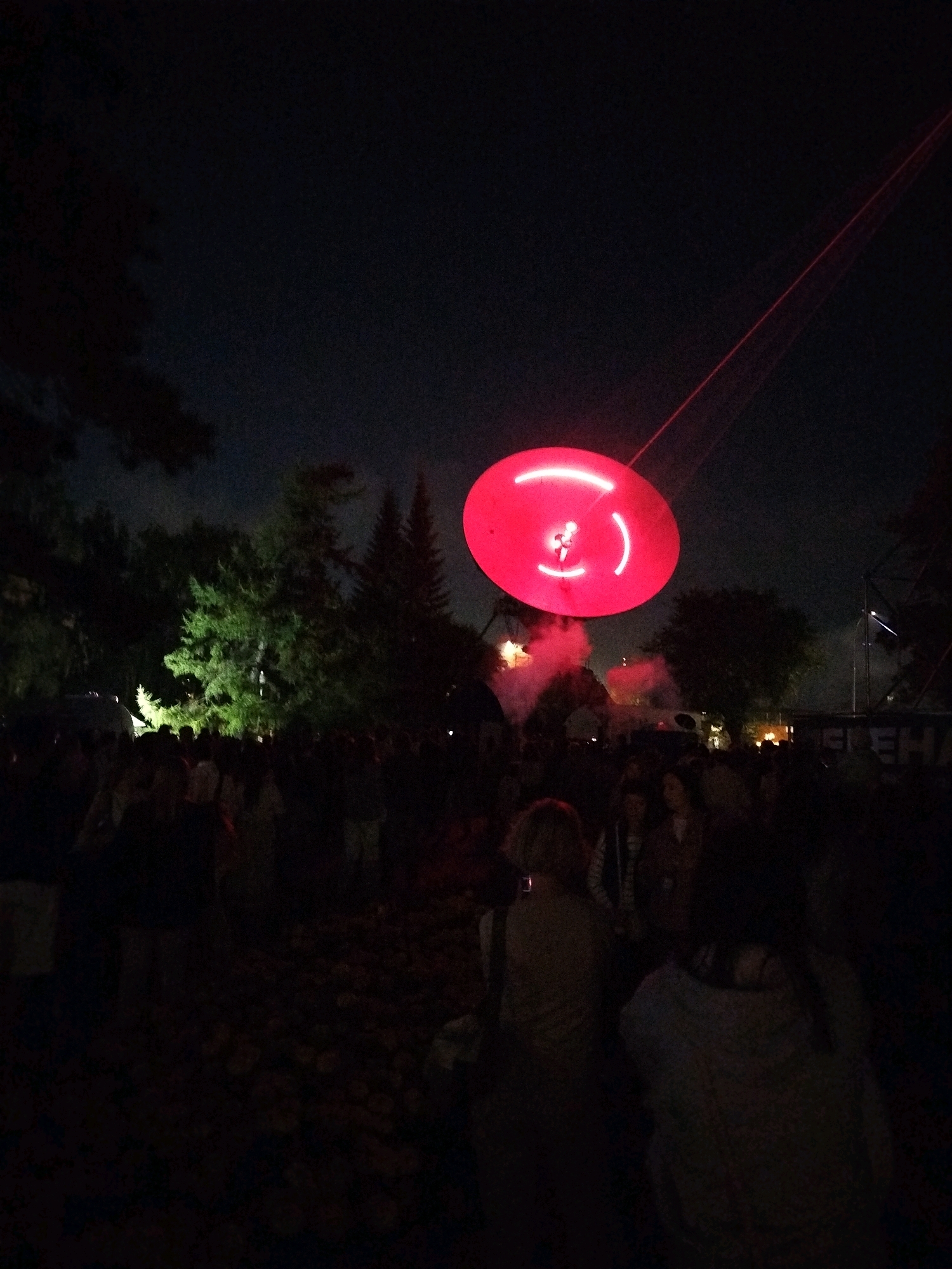 An outdoor evening event is underway in Kemerovo, Russia, with a crowd of people gathered in the foreground. A large, circular, red illuminated object is suspended in the dark sky, emitting a red laser beam that extends upwards. The object has a bright, white, circular outline with smaller circular shapes within. The scene is dimly lit, with the red object and a illuminated tree in the middle background being the primary light sources. The crowd appears to be looking up at the illuminated object, with some individuals holding up phones, possibly to capture images or videos of the spectacle. The environment suggests a park or open space, with trees and some structures visible in the background, including a partially obscured sign.