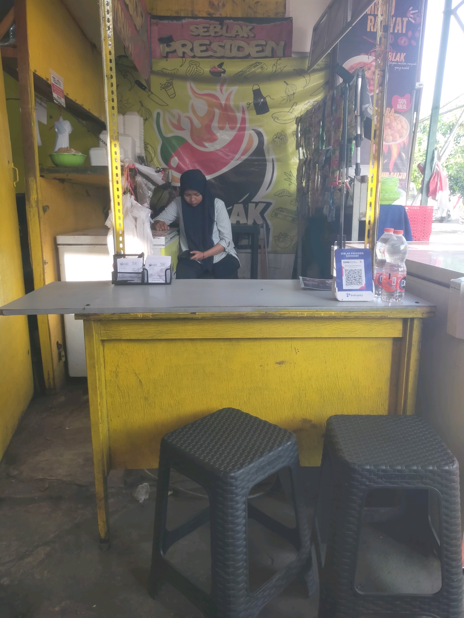 The image shows an outdoor food stall in Sidodadi, Indonesia. A woman wearing a black hijab and a striped shirt is standing behind a yellow counter, looking at her phone. On the counter, there is a QR code payment stand, two bottles of water, and a card holder with some business cards. In front of the counter, there are two black plastic stools. The stall has a yellow wall and a banner with Indonesian text and graphics of food. The overall impression is a small, casual food business.