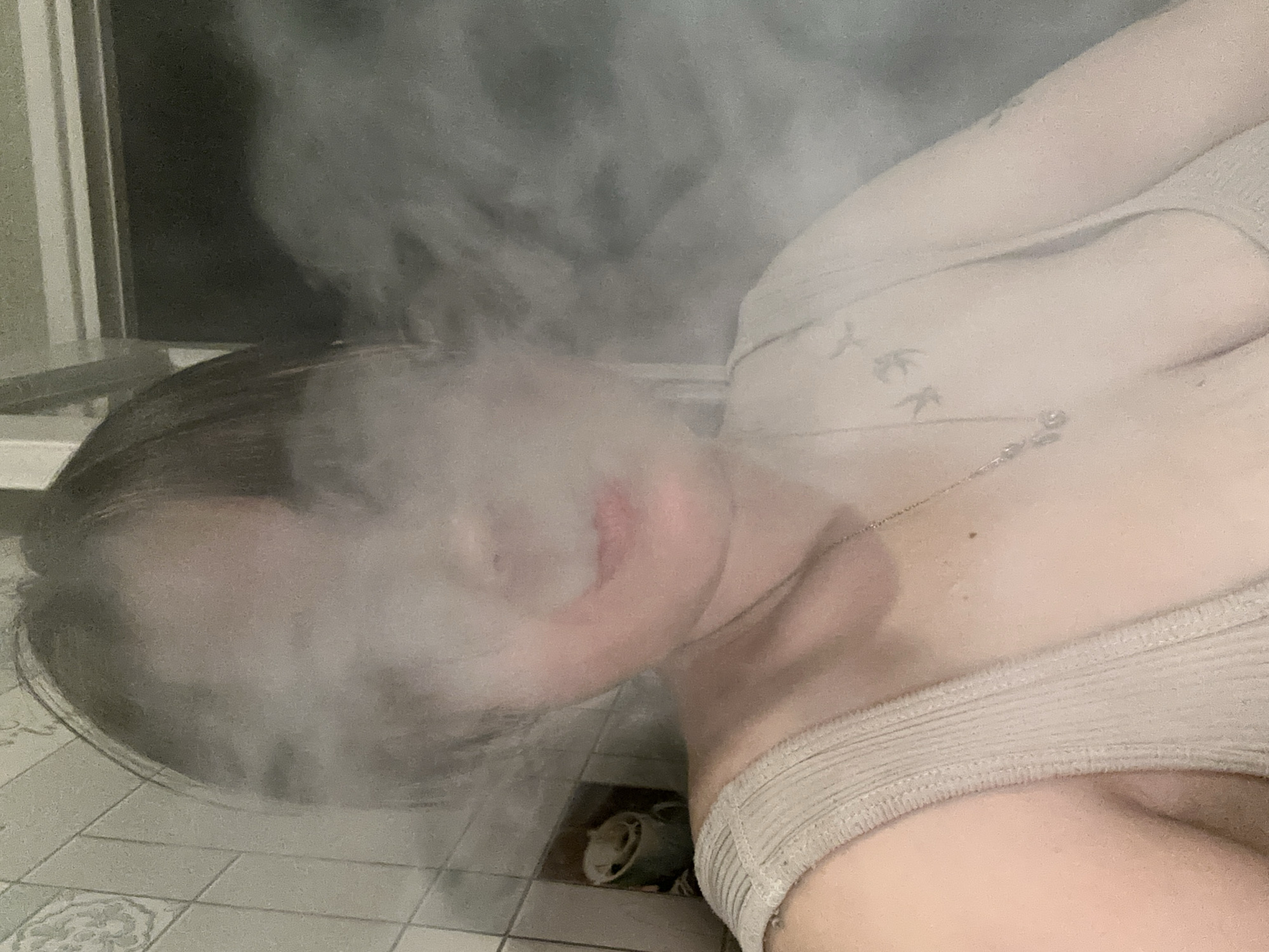 This media file is a close-up, slightly tilted, indoor photograph of a person. The subject is obscured by a dense cloud of white smoke or vapor, with only a portion of their face visible, including red-tinted lips. The person appears to be wearing a light-colored, ribbed tank top and a delicate necklace with a small pendant. A faint tattoo of what appears to be birds in flight is visible on their shoulder. The setting is tiled, with a geometric pattern on the floor. In the background, a dark, indistinct area suggests a wall or window. The overall mood is somewhat mysterious due to the obscuring smoke. There is no visible text in the image.