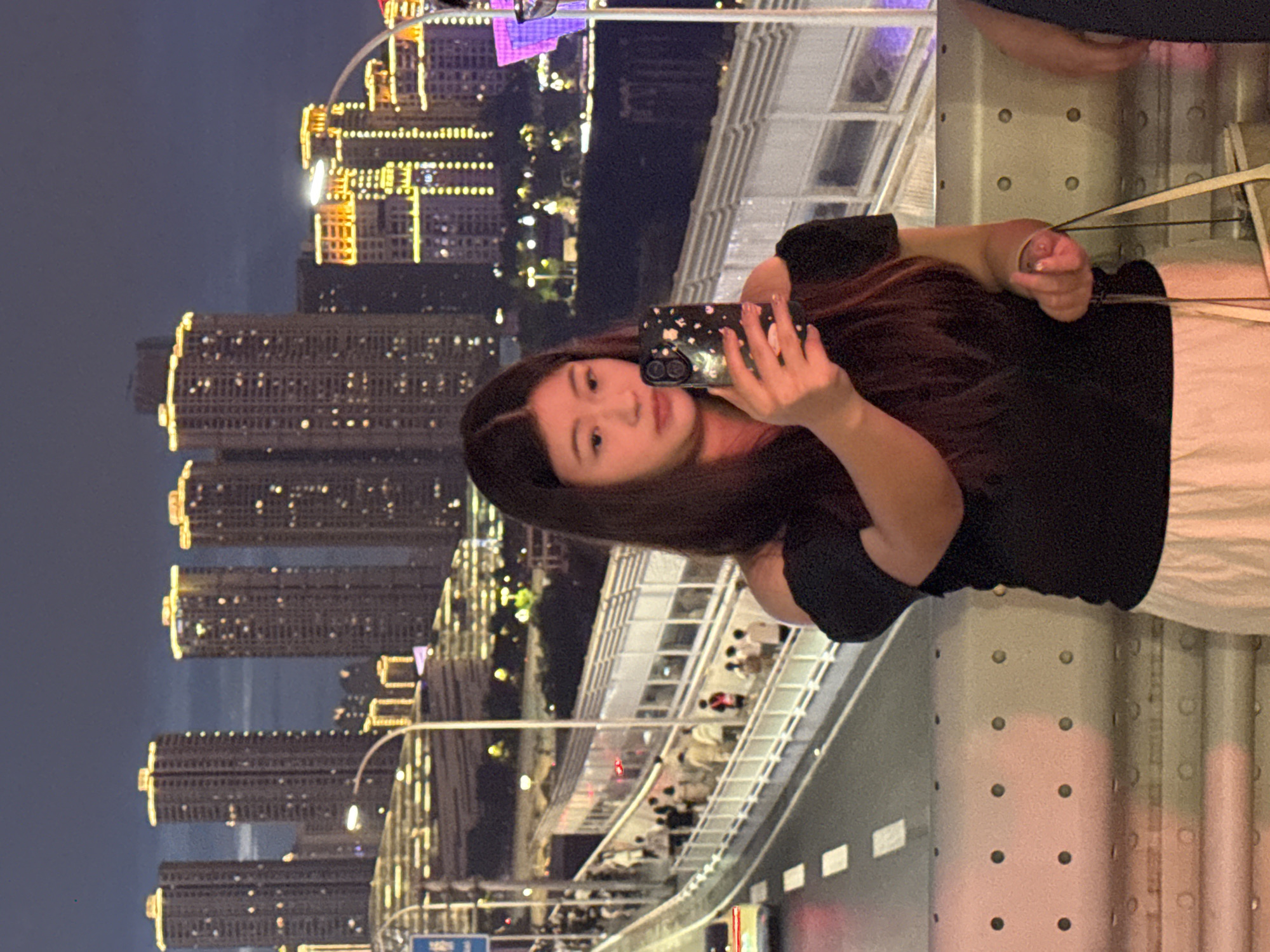 A young East Asian woman is standing on a bridge, taking a selfie with her smartphone. She has long, dark hair and is wearing a black off-the-shoulder top and a white skirt. The background shows a cityscape at dusk, with tall buildings illuminated by numerous lights. The bridge has a pedestrian walkway and a road with a vehicle visible. The location is identified as Yuzhong District, China. The overall scene is nocturnal, with a clear sky above the city lights.