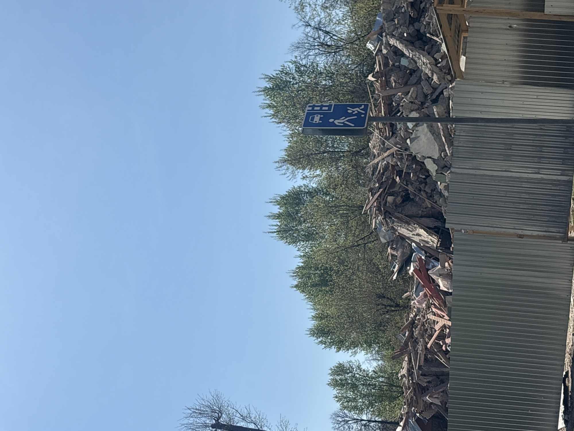 The image shows a street scene with a bus stop sign in Perm, Russia. The sign depicts a bus and pedestrians. Behind the sign, there is a large pile of rubble and debris, possibly from a construction or demolition site. A corrugated metal fence is adjacent to the debris. The sky is clear and blue. Some trees are visible in the background.