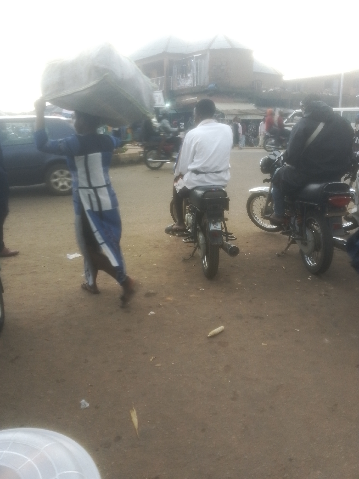 This media file captures a bustling street scene in Gwong, Nigeria. The main subject is a person walking from left to right, carrying a large, light-colored bundle balanced on their head. They are dressed in a blue and white patterned garment. Behind this person, two individuals are seated on motorcycles. The person on the left is wearing a white shirt, while the person on the right is wearing a black jacket. The motorcycles appear to be typical of those used for transportation in the region.
The setting is an outdoor urban environment, likely a market or busy thoroughfare. Buildings with corrugated iron roofs and balconies are visible in the background, suggesting a commercial area. The ground is unpaved, a dusty dirt surface. The lighting indicates it is daytime, possibly late afternoon, with a slightly overcast sky. The overall atmosphere suggests everyday life and commerce, with people engaged in their daily activities. There are no clearly visible signs or text in the image. The focus on the individuals and their activities, along with the surrounding environment, provides a glimpse into local life in Gwong.