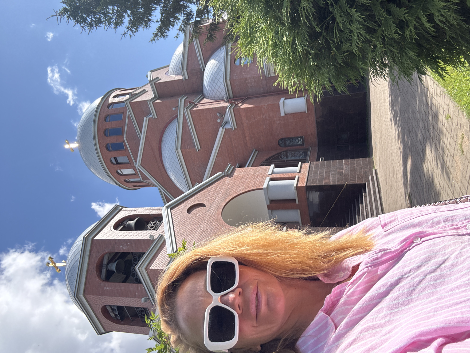 A woman wearing large white sunglasses and a pink striped collared shirt is taking a selfie in front of a red brick Orthodox church with multiple silver domes. The church has a bell tower with bells visible. The sky is blue with some white clouds. Lush green trees and bushes are present on the right side of the image. The ground in front of the church is paved with interlocking bricks. The image appears to have been taken on a sunny day. The woman's hair is long and blonde.