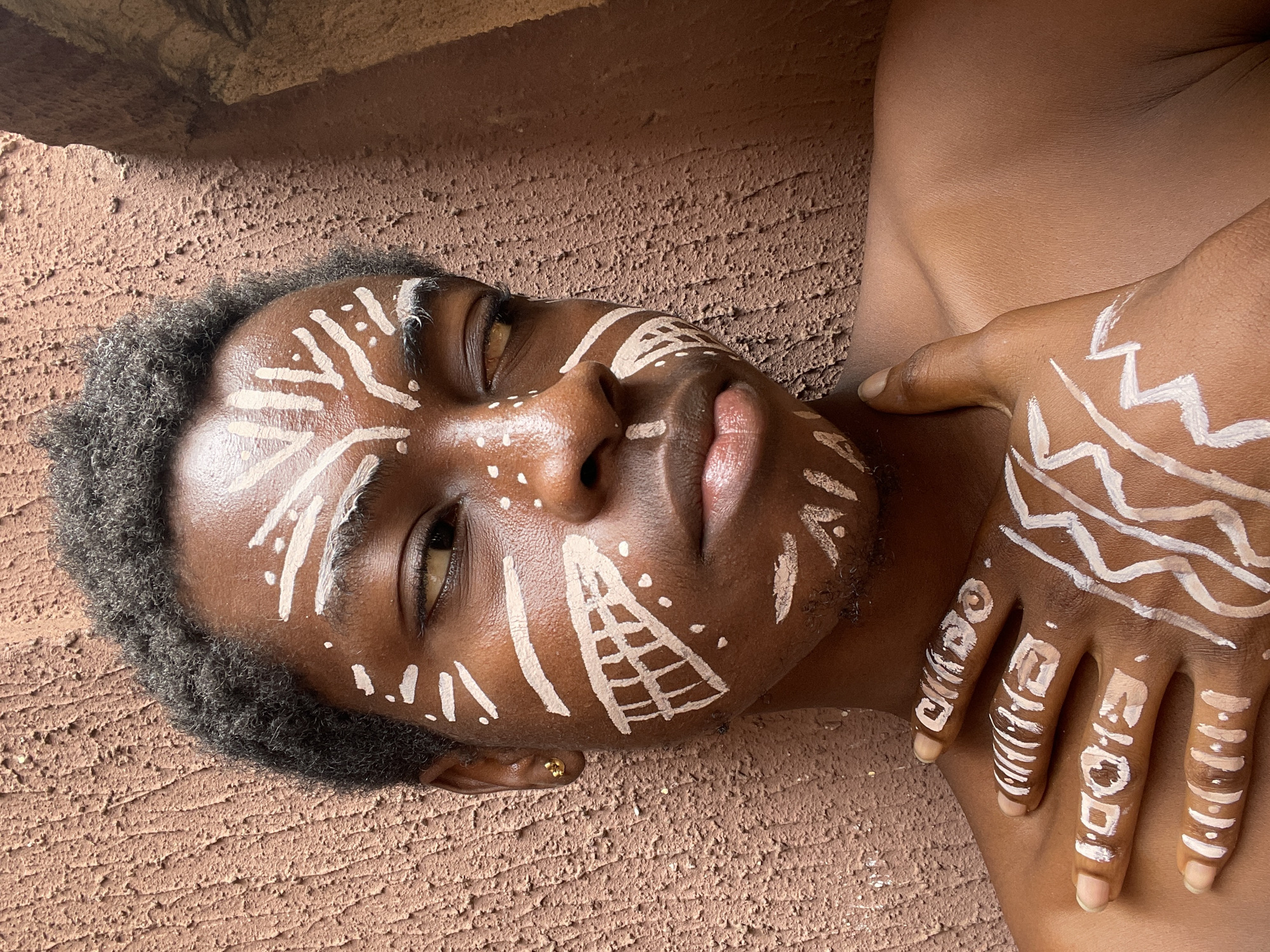 The image displays a person with dark skin, adorned with intricate white body paint on their face and left hand, positioned against a textured, reddish-brown wall. The subject's head is tilted upwards, with eyes partially open and gazing off-camera, conveying a serene or contemplative expression. Short, dark, curly hair frames the face, which also exhibits light stubble. A small gold earring is visible in the left earlobe.
The white body paint consists of diverse abstract patterns. On the face, these include lines, dots, zigzag motifs, and a prominent conical or pyramid-like design with internal cross-hatching on the right cheek. Similar markings are present on the forehead, left cheek, and chin. The left hand, resting gently across the chest or shoulder, is also extensively covered in white paint, featuring zigzag patterns on the back. On the fingers, stylized patterns are visible; the middle finger clearly displays a stylized word resembling "BLOOD," and the index finger has other distinct symbols or letters. The subject's fingernails are natural and unpolished.
The background is a rough, uneven, reddish-brown surface, consistent with a textured wall, providing a stark contrast to the white body art. The lighting is bright and even, suggesting a daytime setting, possibly indoors or in a shaded outdoor area. The overall scene captures a moment of cultural expression or artistic posing within what appears to be a natural or traditional environment, given the location context of Nkpor, Nigeria.