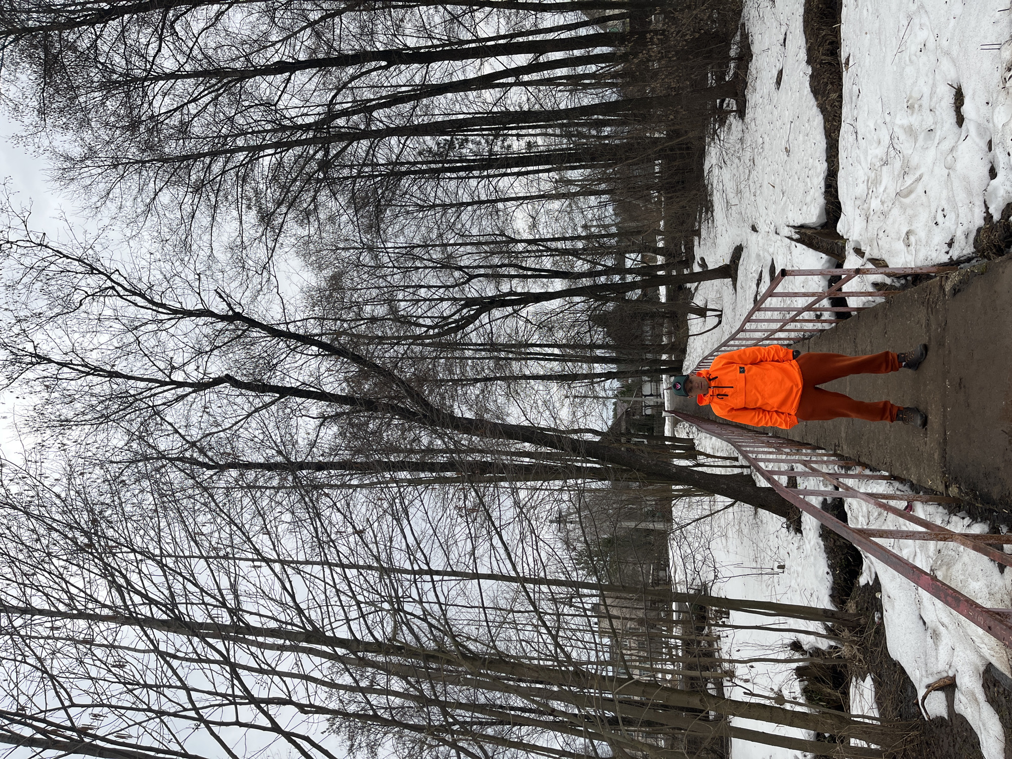 A person in an orange jacket and pants is standing on a concrete structure with metal railings in what appears to be a park or wooded area in Chaikovsky, Russia. Patches of snow are visible on the ground. The trees are bare, suggesting it is winter or early spring. The sky is overcast.